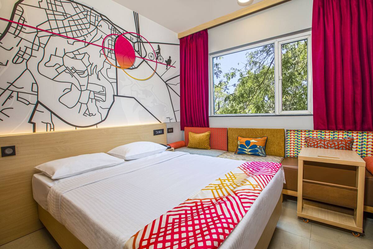 Una habitación de hotel moderna con una cama grande, un mural de colores vivos, una ventana con cortinas rojas y una pequeña zona de estar con cojines.