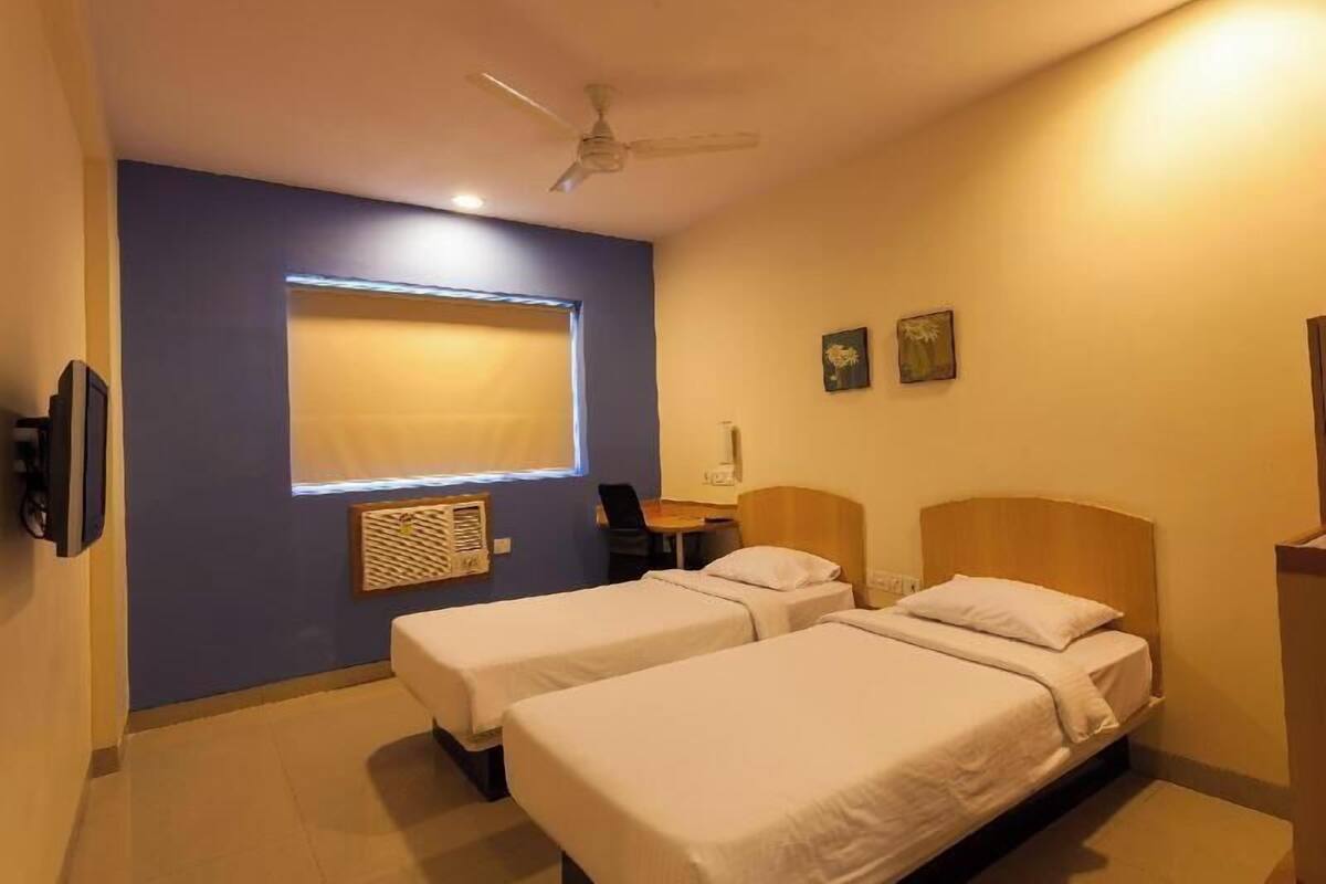 Habitación de hotel con dos camas individuales, ventilador de techo, ventana con persianas, escritorio y una silla.