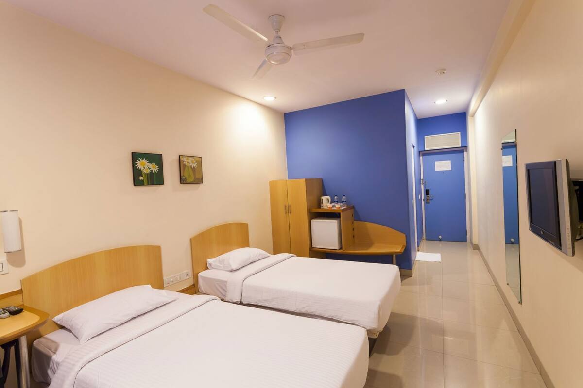 A hotel room with two beds, a desk, a wardrobe, a television, and a ceiling fan.