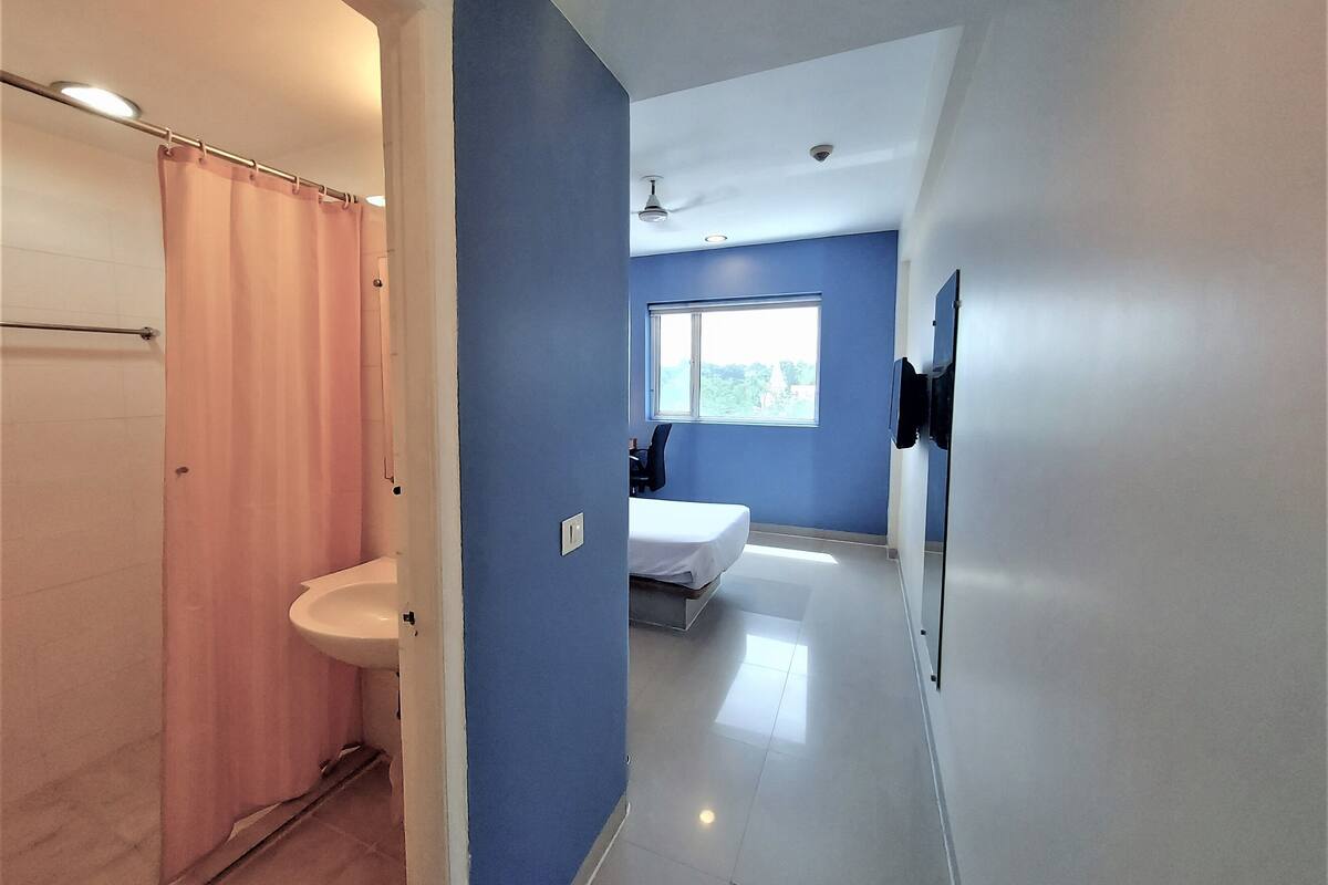 Un baño moderno con ducha, inodoro y lavamanos. En la habitación contigua hay una cama y un ventanal que proporciona luz natural.