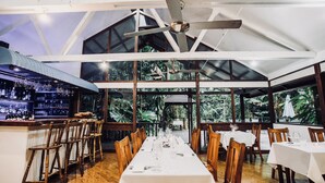 A restaurant with a long table set for dining, wooden chairs, and a bar area with bottles. - Daintree Wilderness Lodge (Diwan)