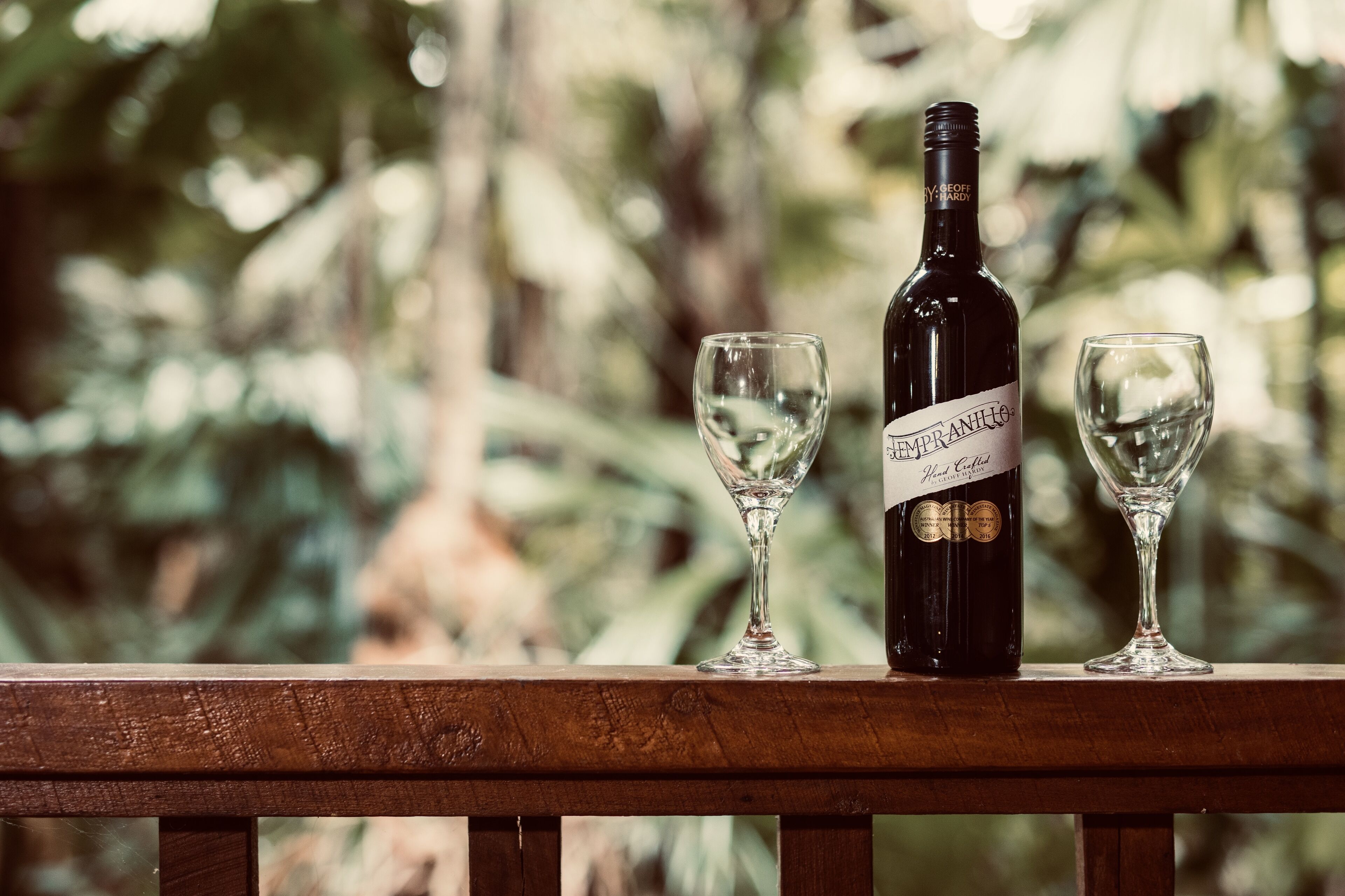 A wine bottle and two glasses on a wooden railing with a blurred natural background.