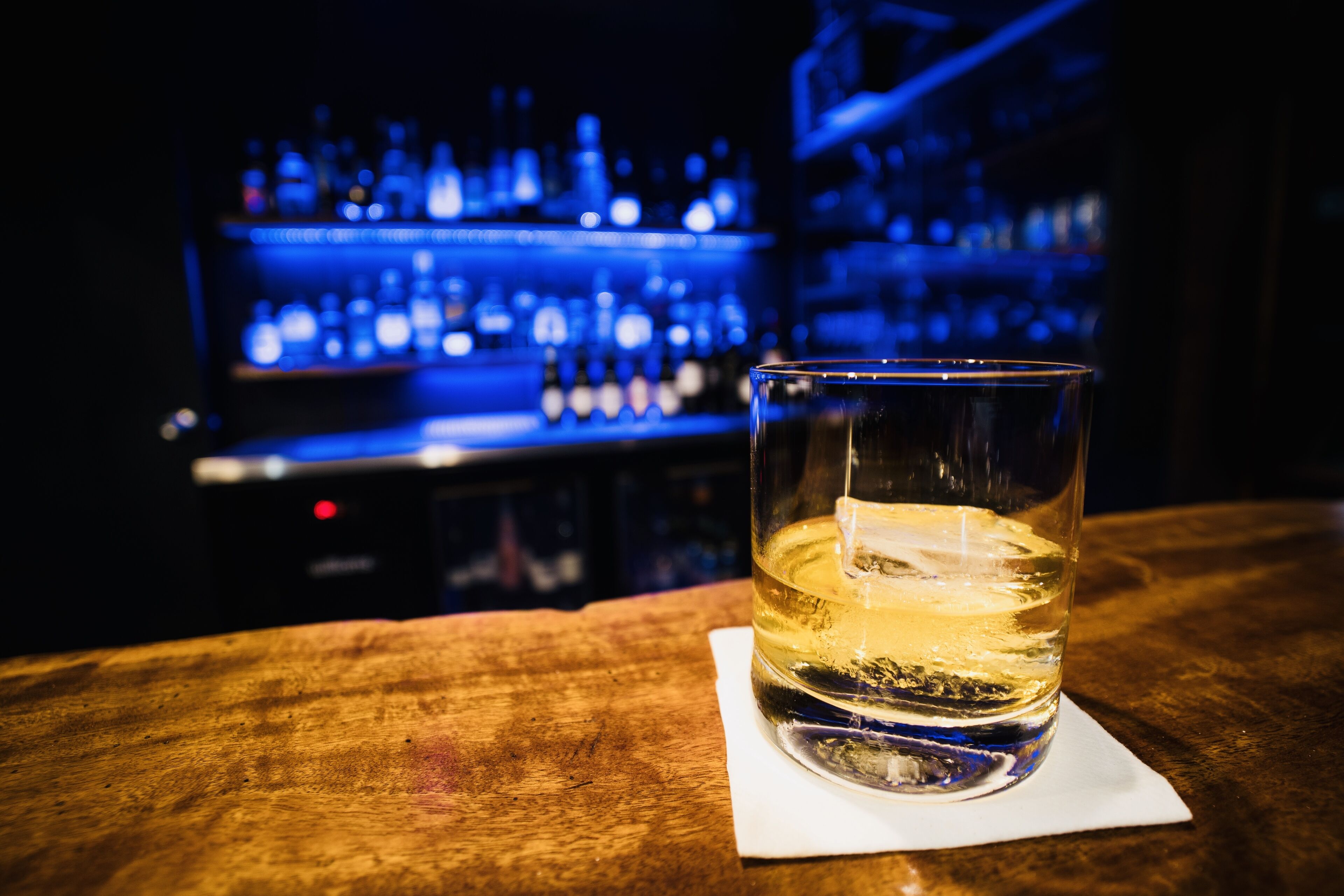 A glass of whiskey on a wooden bar counter with a blurred background of shelves stocked with bottles.
