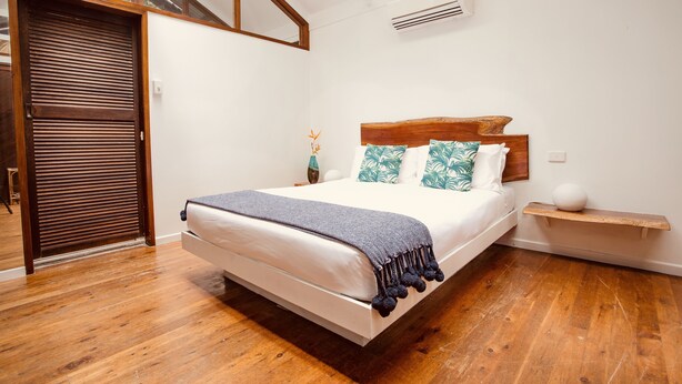Un dormitorio con suelo de madera, una cama con colchón blanco y manta oscura, cabecero de madera y una mesita de noche con un jarrón.