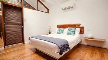 A bedroom with a wooden floor, a bed with a white mattress and a dark blanket, wooden headboard, and a small bedside table with a vase.