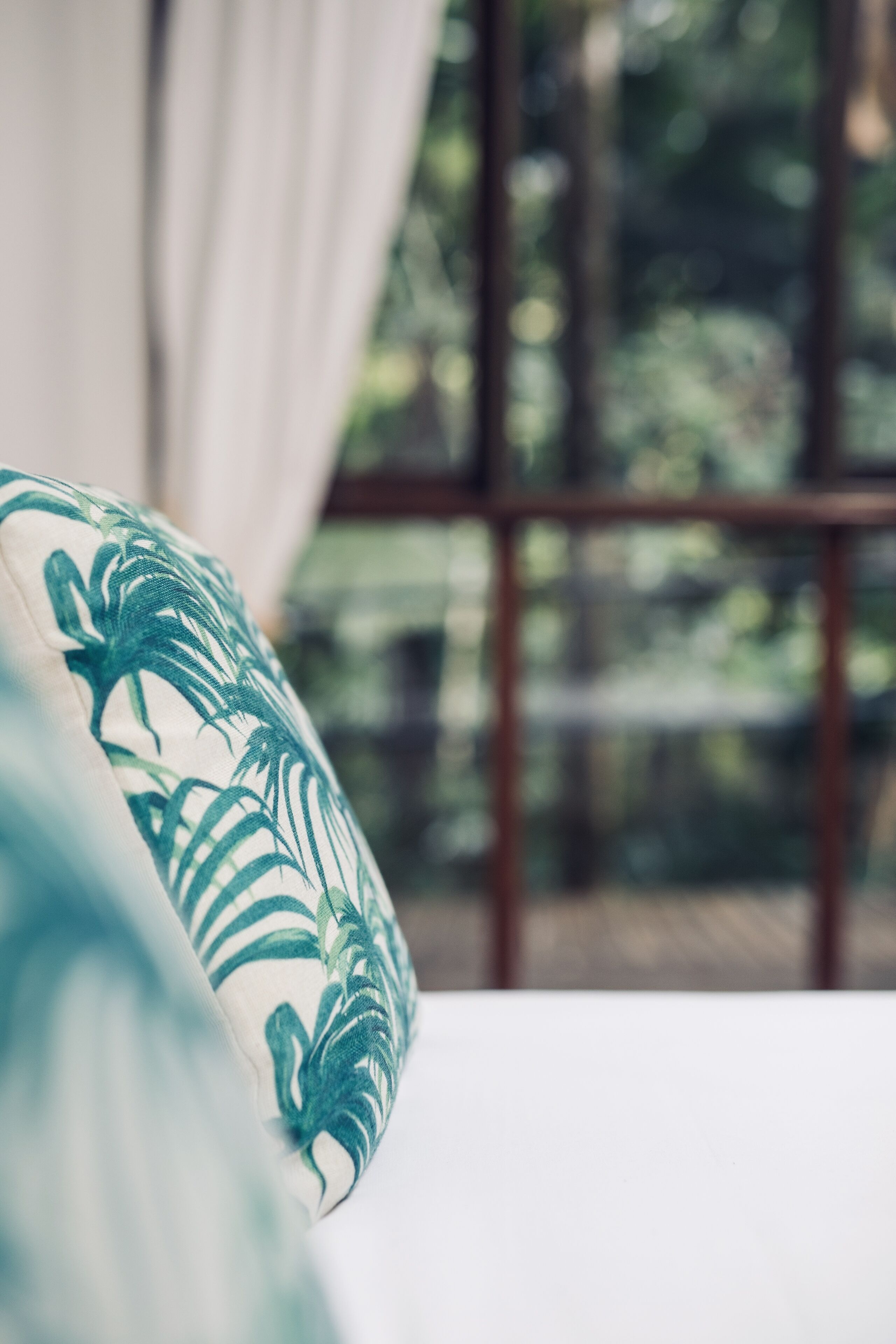 Primer plano de una almohada con un estampado de hojas tropicales, una ventana que da a vista de árboles y una cortina entreabierta.