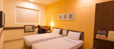 A hotel room with two beds, a desk with a chair, a flat-screen TV, a ceiling fan, and a wall-mounted shelf with bottled water and a coffee maker.
