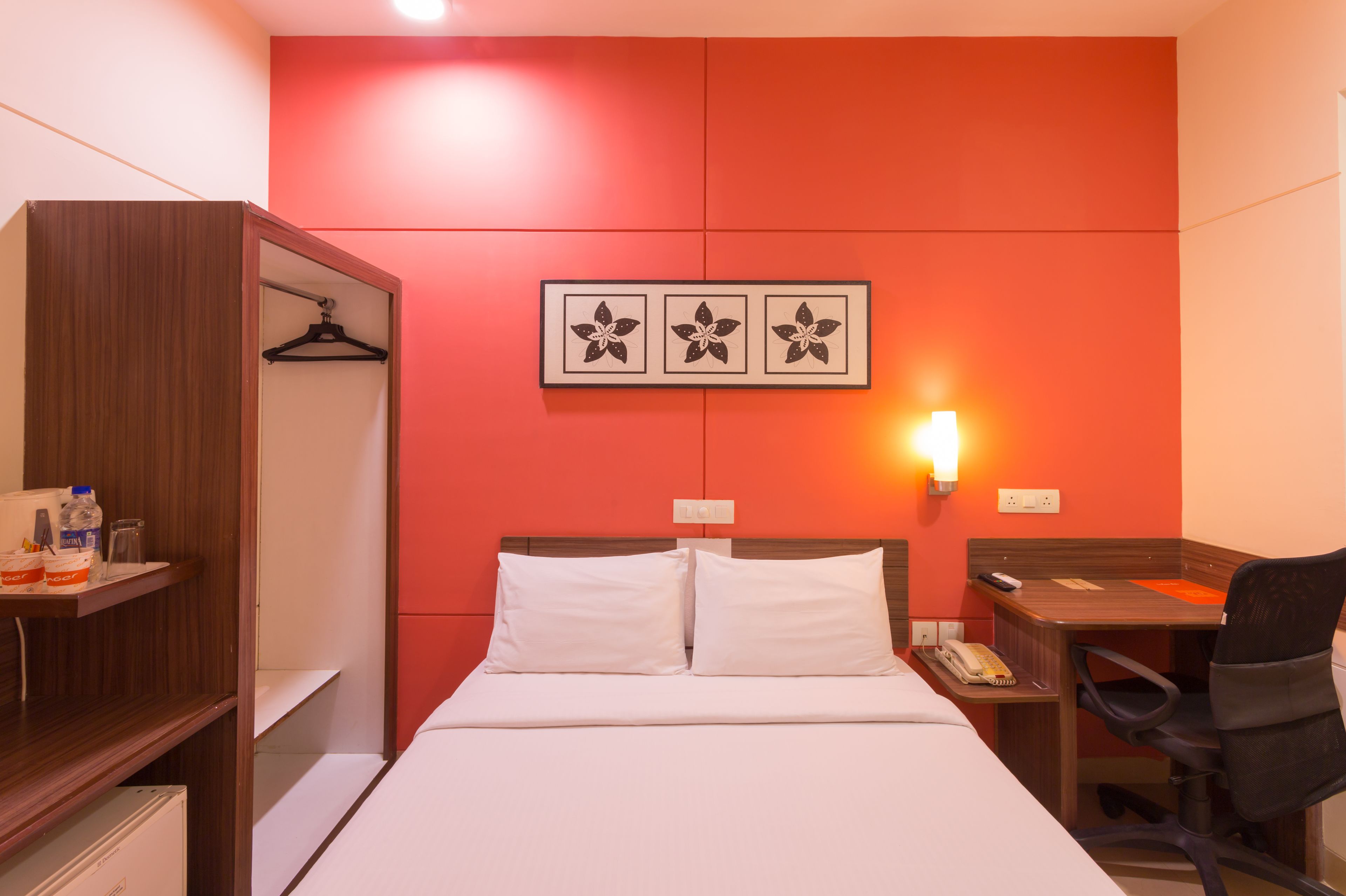 A hotel room with a bed, a desk, a chair, a wardrobe, and a wall-mounted light.