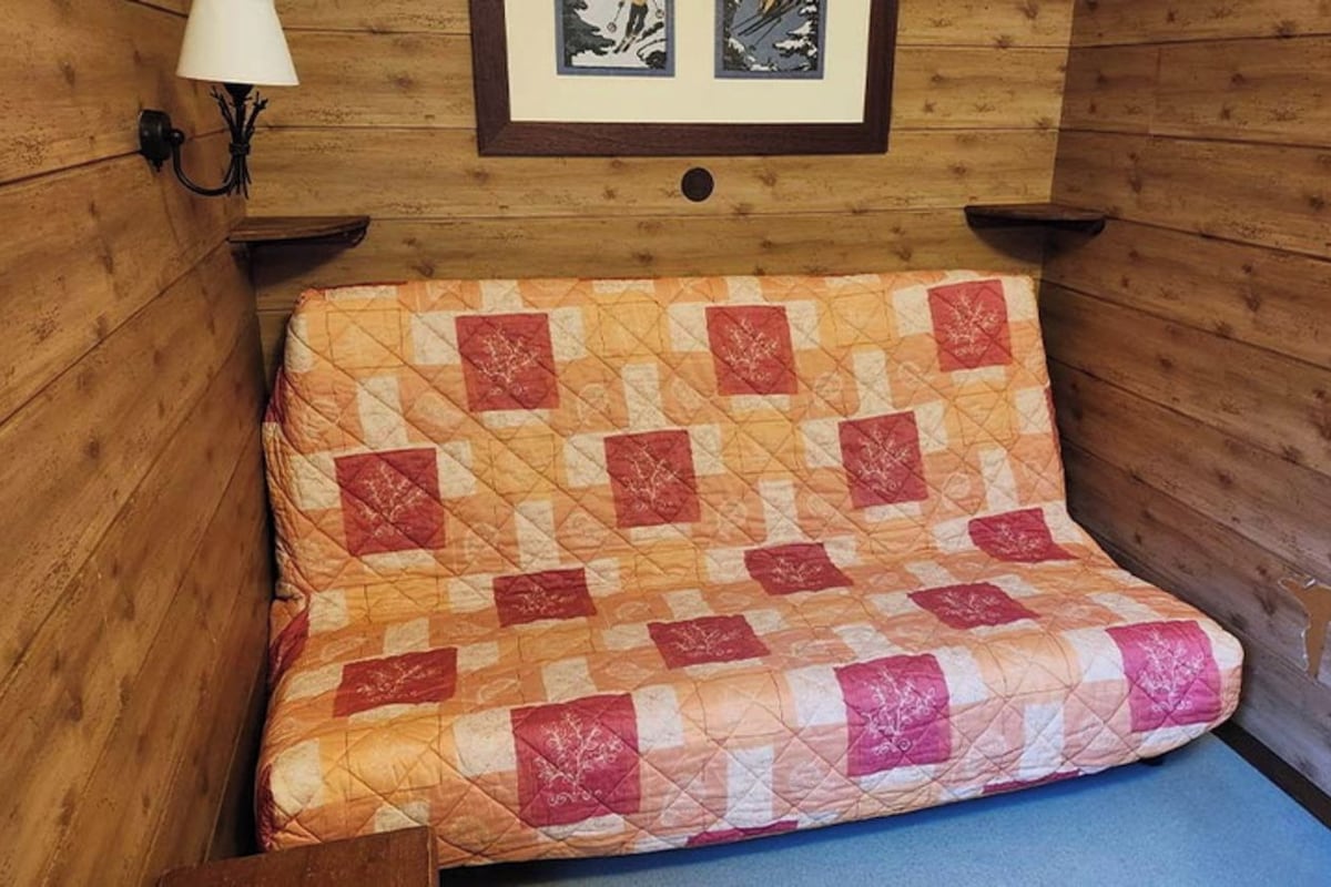 L’intérieur d’un chalet en bois, avec un lit recouvert d’une courtepointe, une lampe fixée au mur et des tableaux encadrés au-dessus du lit.