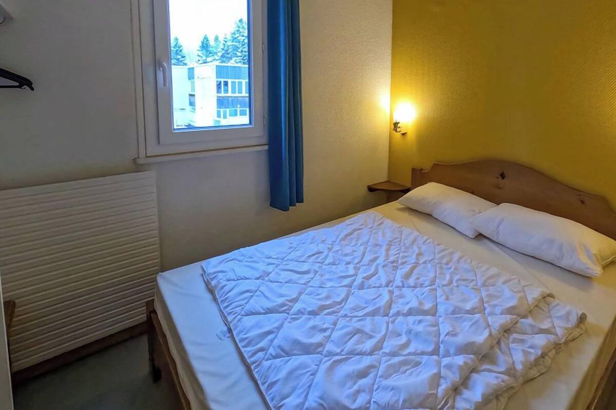 A hotel room with a bed, a window with curtains, a wall-mounted light, and a radiator.