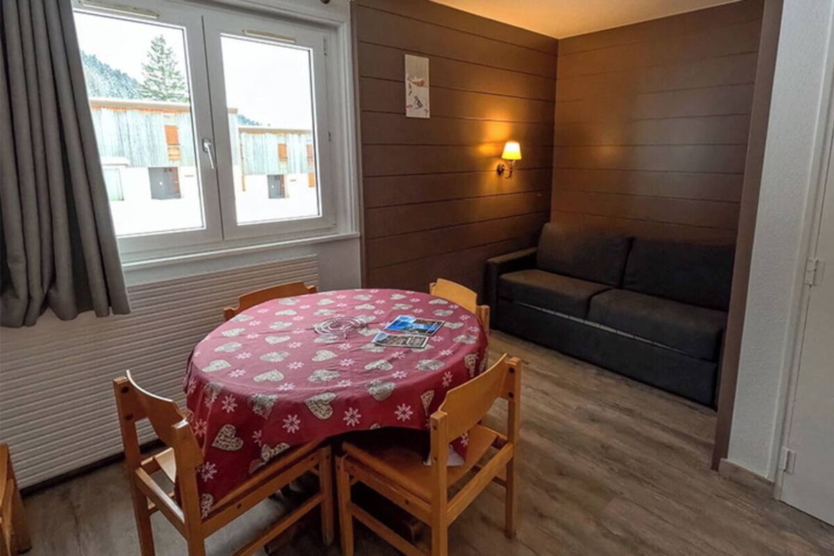 A room with a round table covered with a red tablecloth, a sofa, a wall-mounted lamp, and a window with curtains.