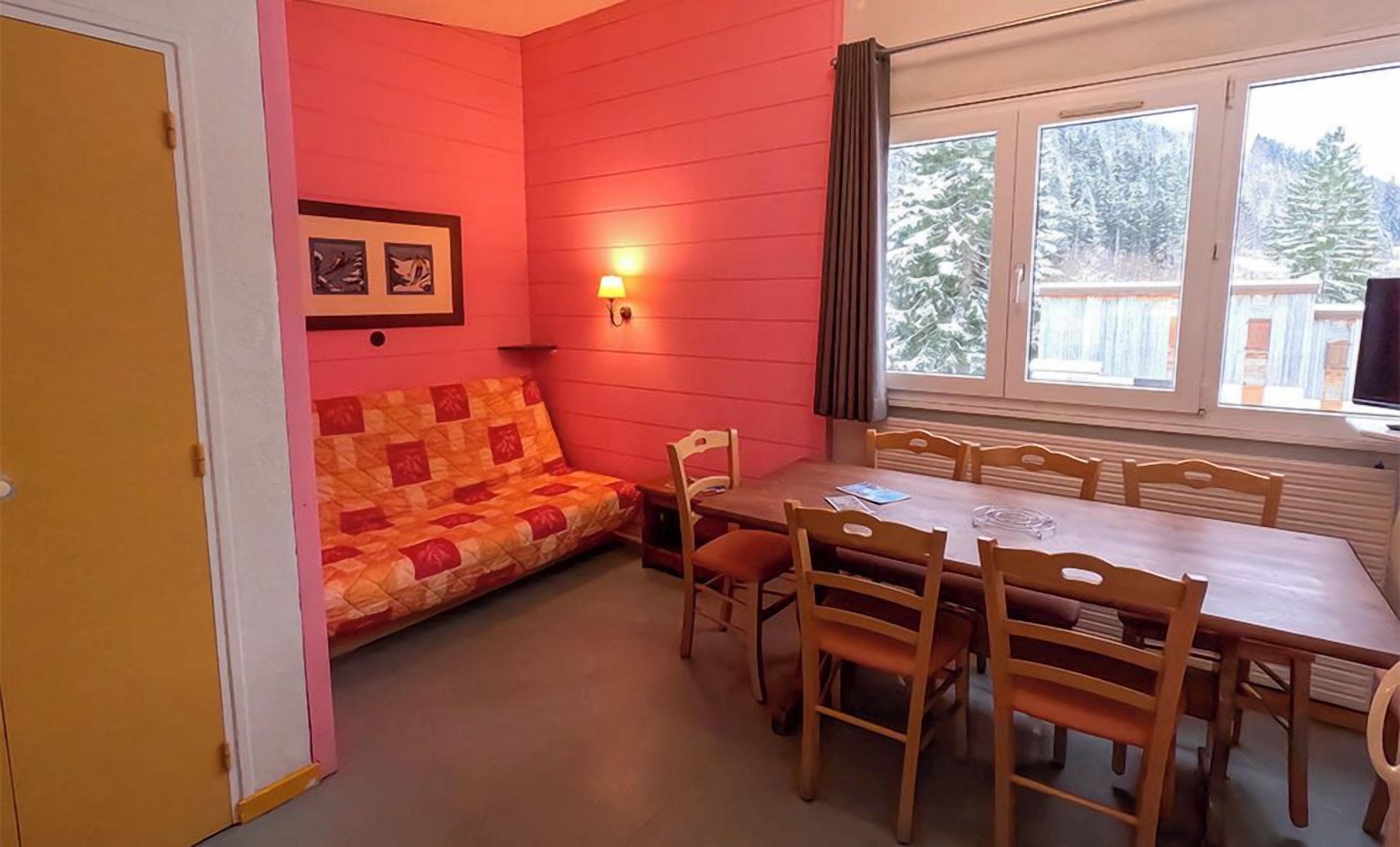 Une pièce avec une table et des chaises en bois, un mur rouge, une fenêtre donnant sur des arbres et une porte.