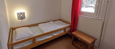 A small, single-bed room with a wooden frame, white bedding, a small wooden bench, a radiator, and a window with red curtains.
