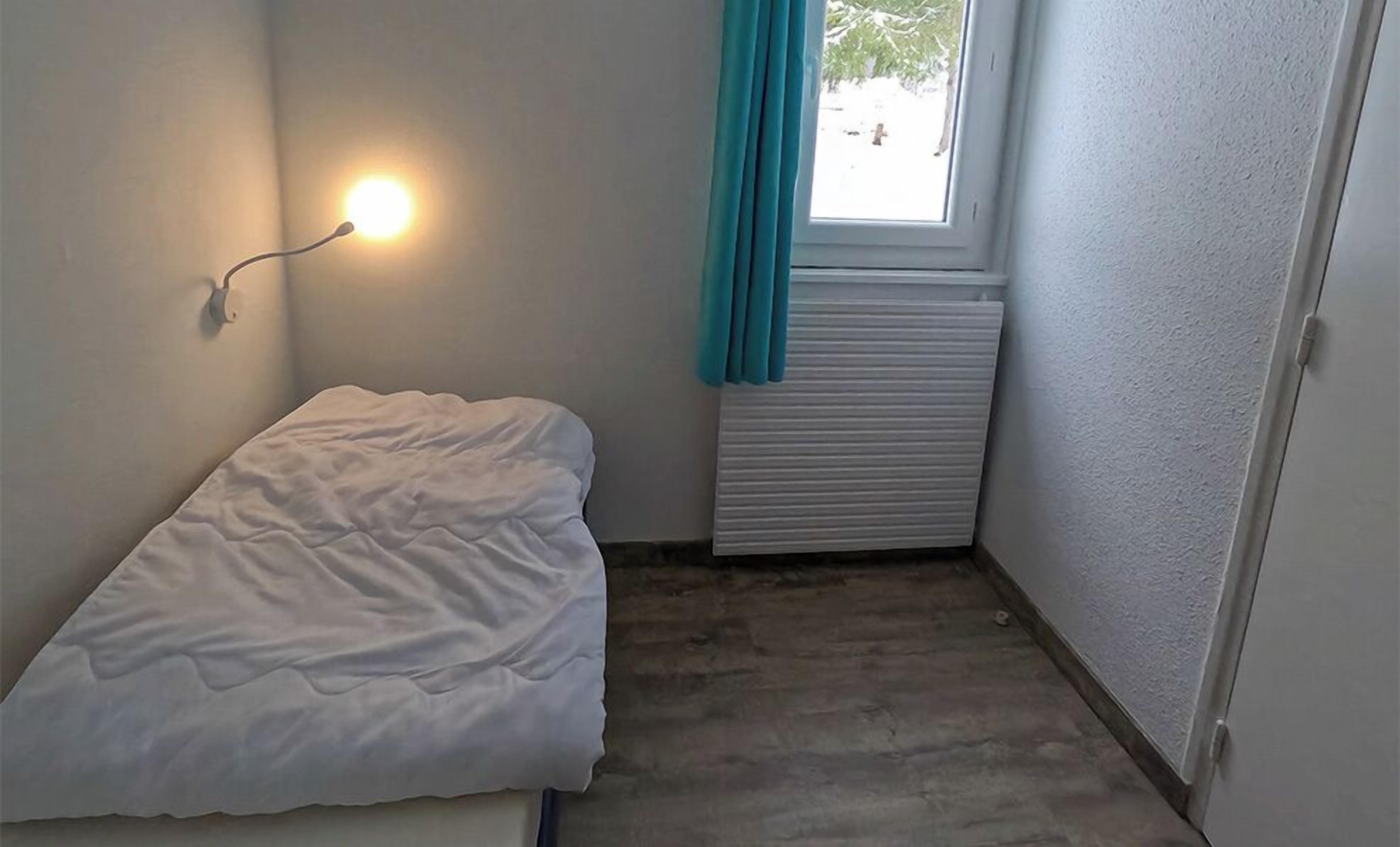 Una habitación pequeña y poco amueblada con una cama individual, una lámpara en la pared, una ventana con cortinas y un radiador.