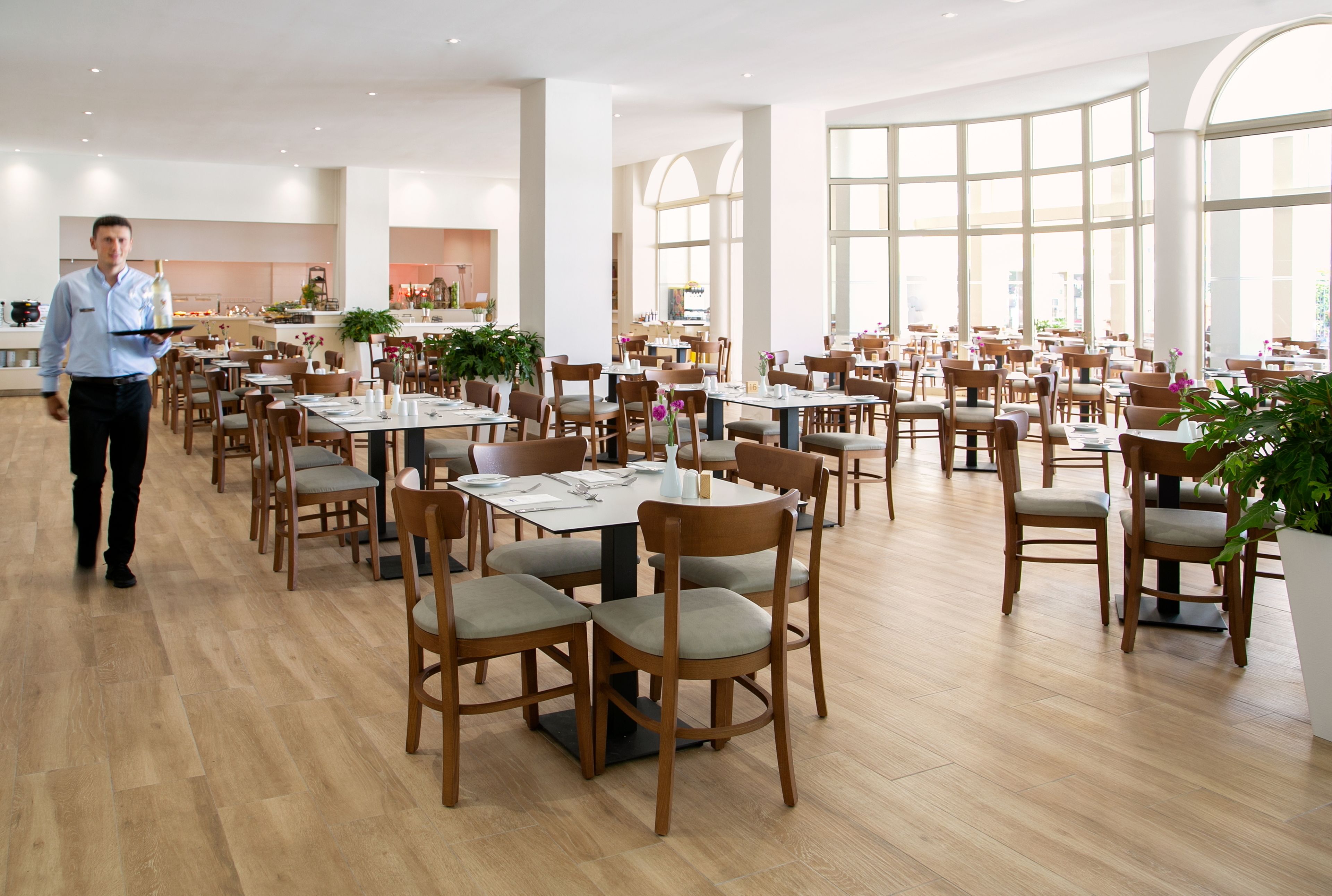 A spacious dining area with wooden tables and chairs, large windows, and a waiter walking.