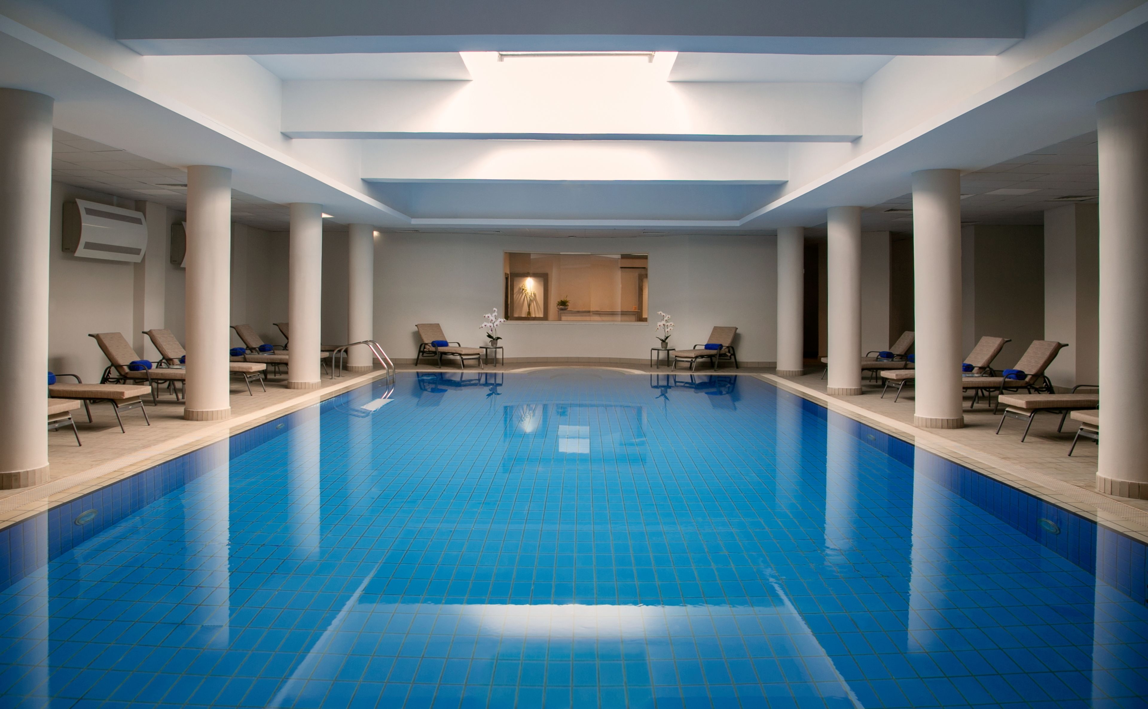 Una piscina interior con baldosas azules cristalinas, rodeada de sillas de descanso y columnas.