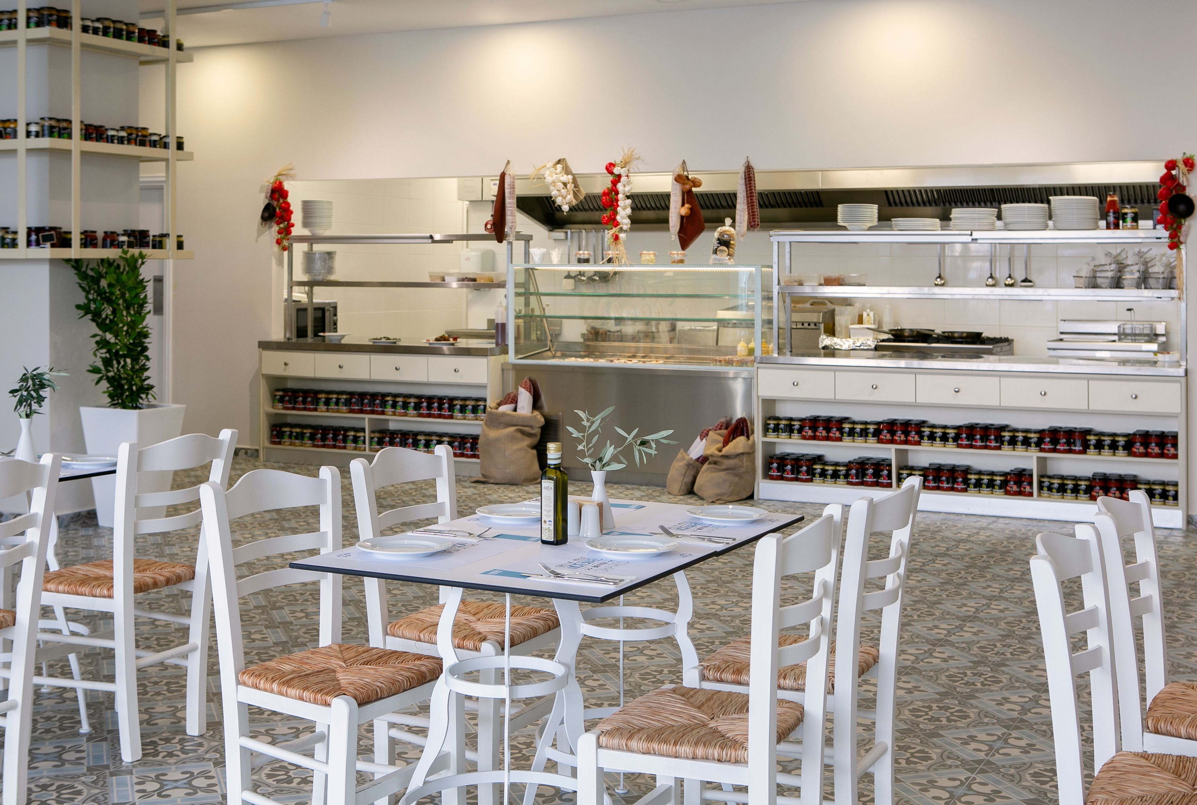A restaurant with white chairs and tables, a glass display case, and shelves with condiments.