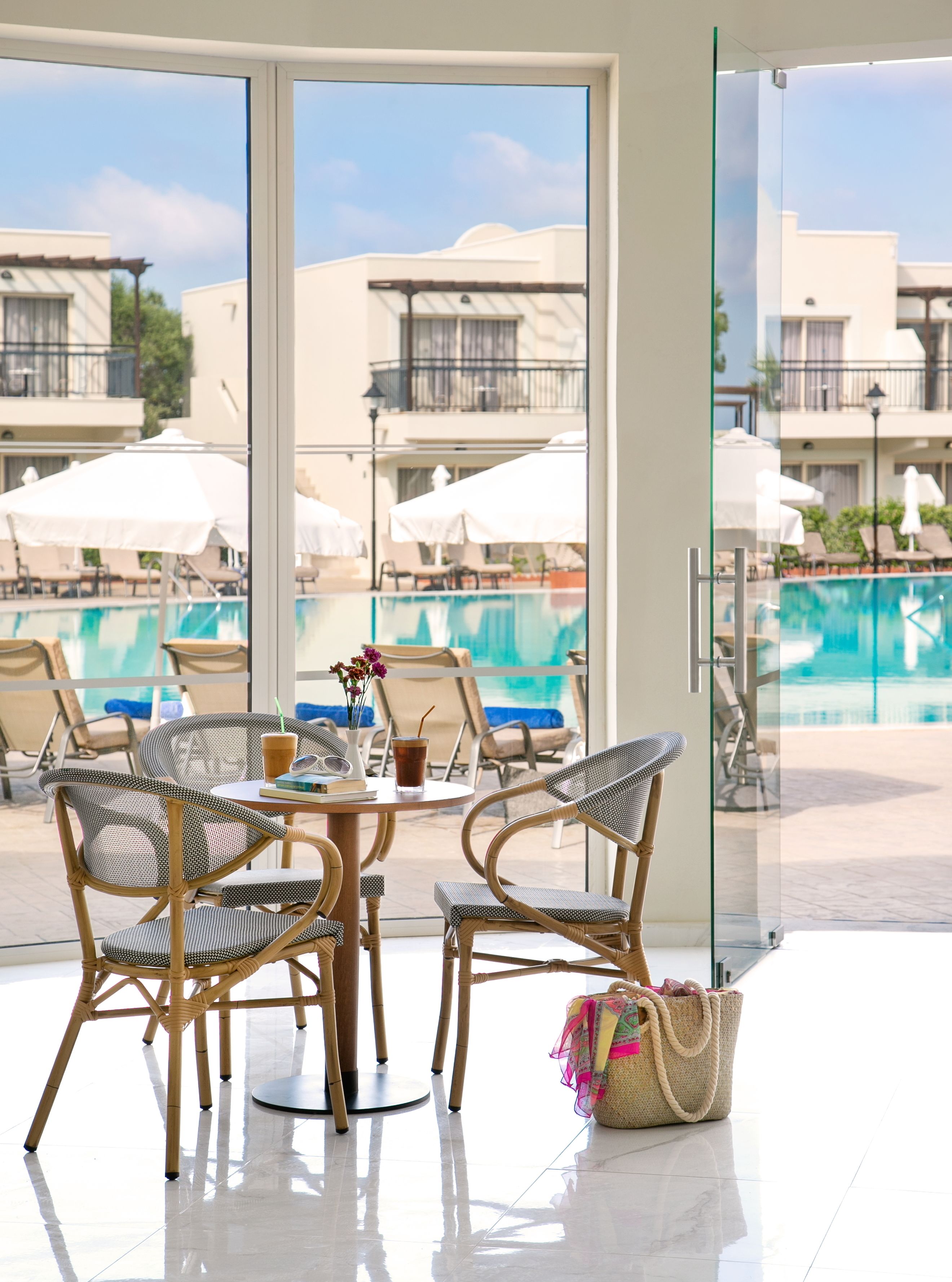 A modern hotel room with a glass door leading to a pool area. There are wicker chairs and a table with a drink and a book.