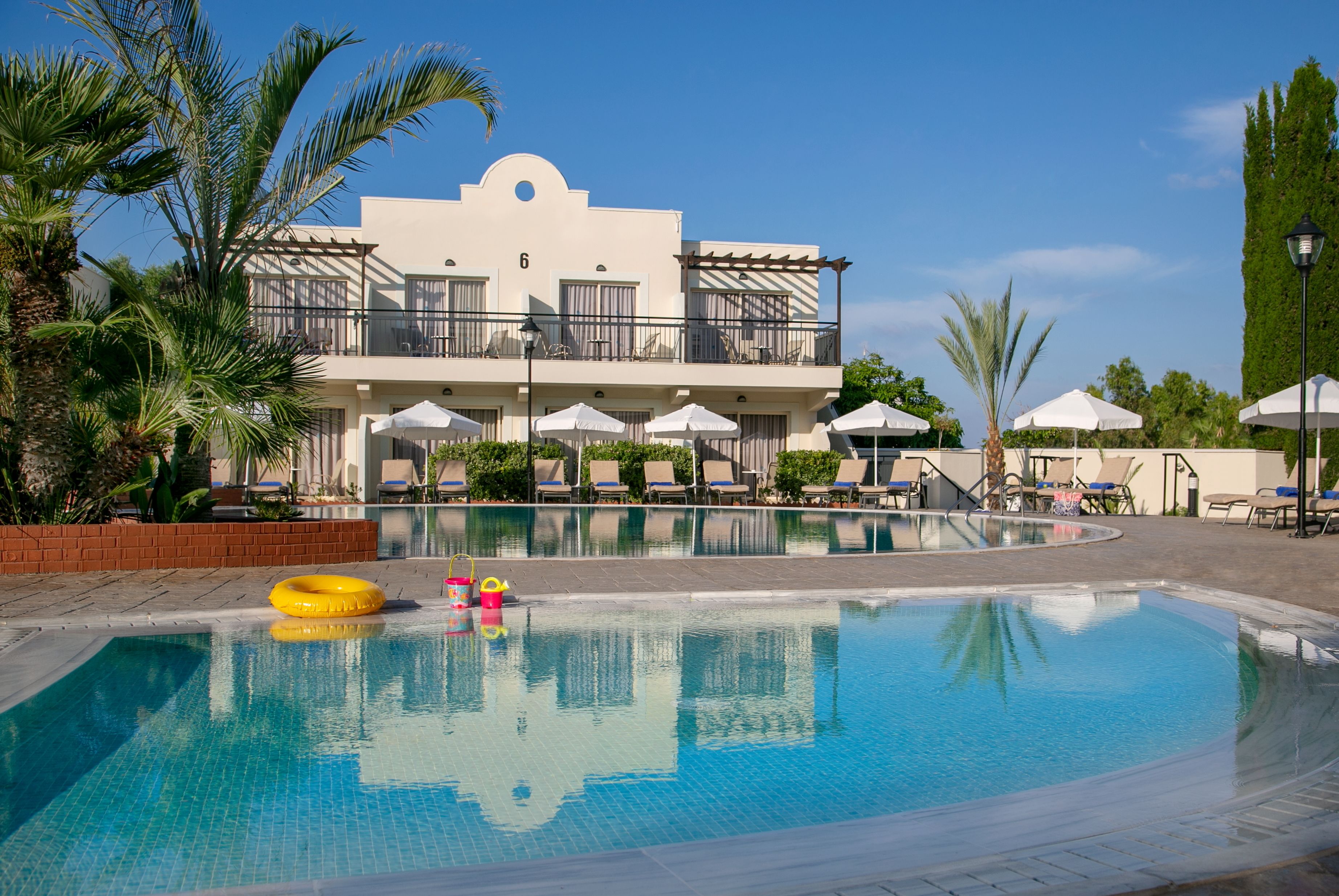Ein Schwimmbecken mit Liegestühlen und Sonnenschirmen, ein Gebäude mit Balkonen und Palmen.