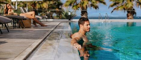 A man in a swimming pool with lounge chairs and umbrellas in the background.