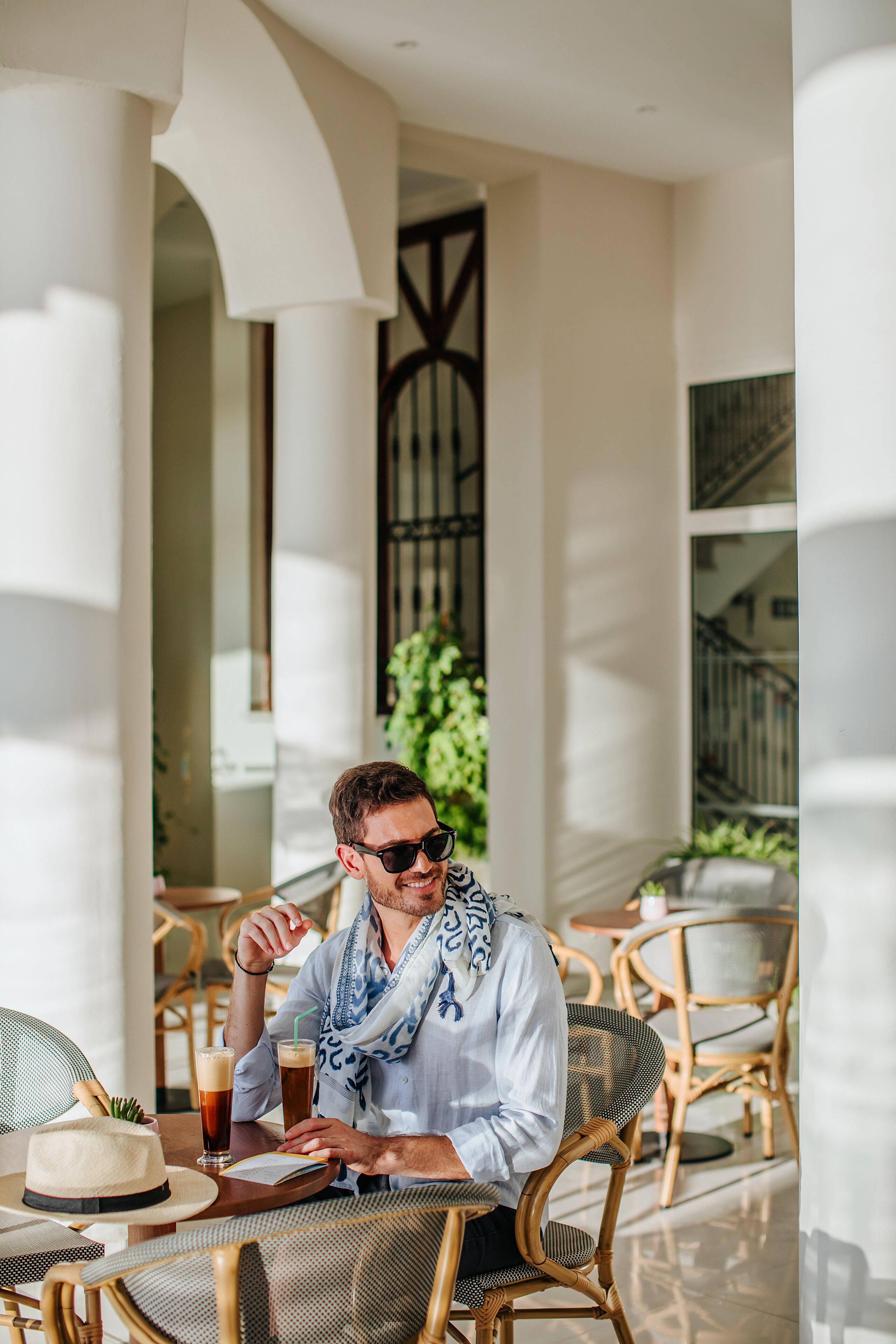 Un homme est assis à une table à l’extérieur, avec une boisson et un chapeau, entouré de chaises en osier et d’une grande fenêtre.