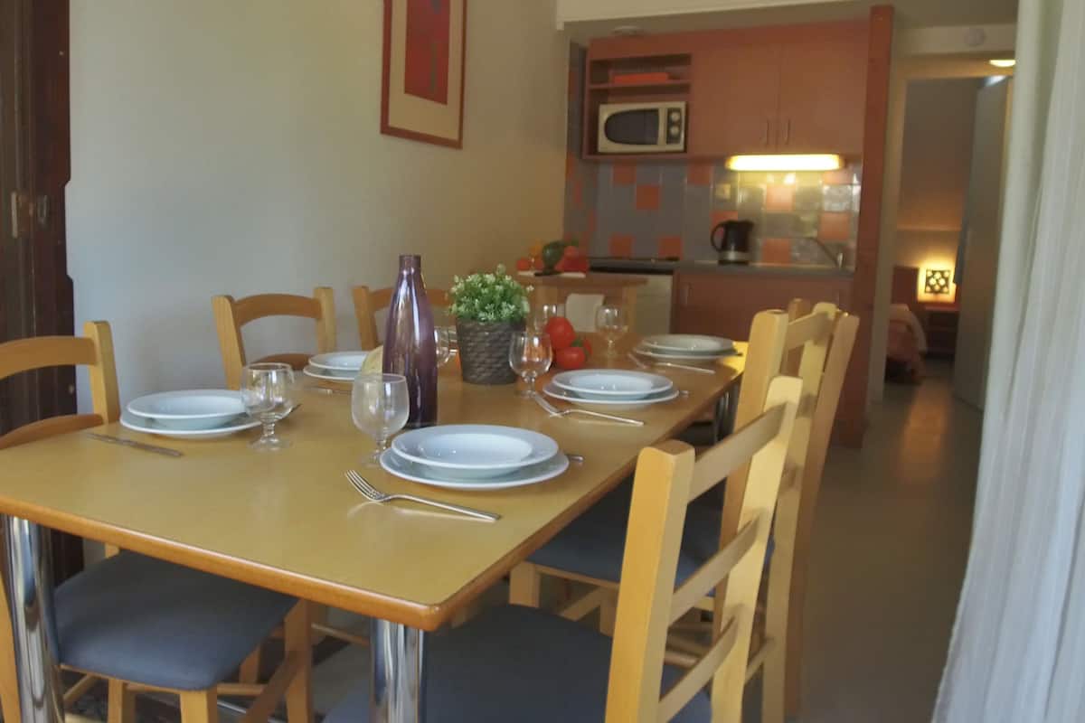 Uma sala de jantar com uma mesa de madeira posta para quatro pessoas, rodeada por cadeiras. Há um vaso com flores e uma fruteira sobre a mesa.