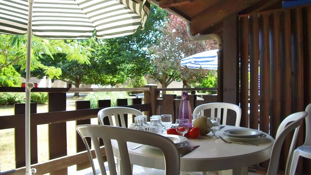 A round table with white chairs, set for dining, under striped umbrellas, with a view of greenery and a building in the background. - VVF Dordogne Lot, Martel-Gluges (Montvalent)