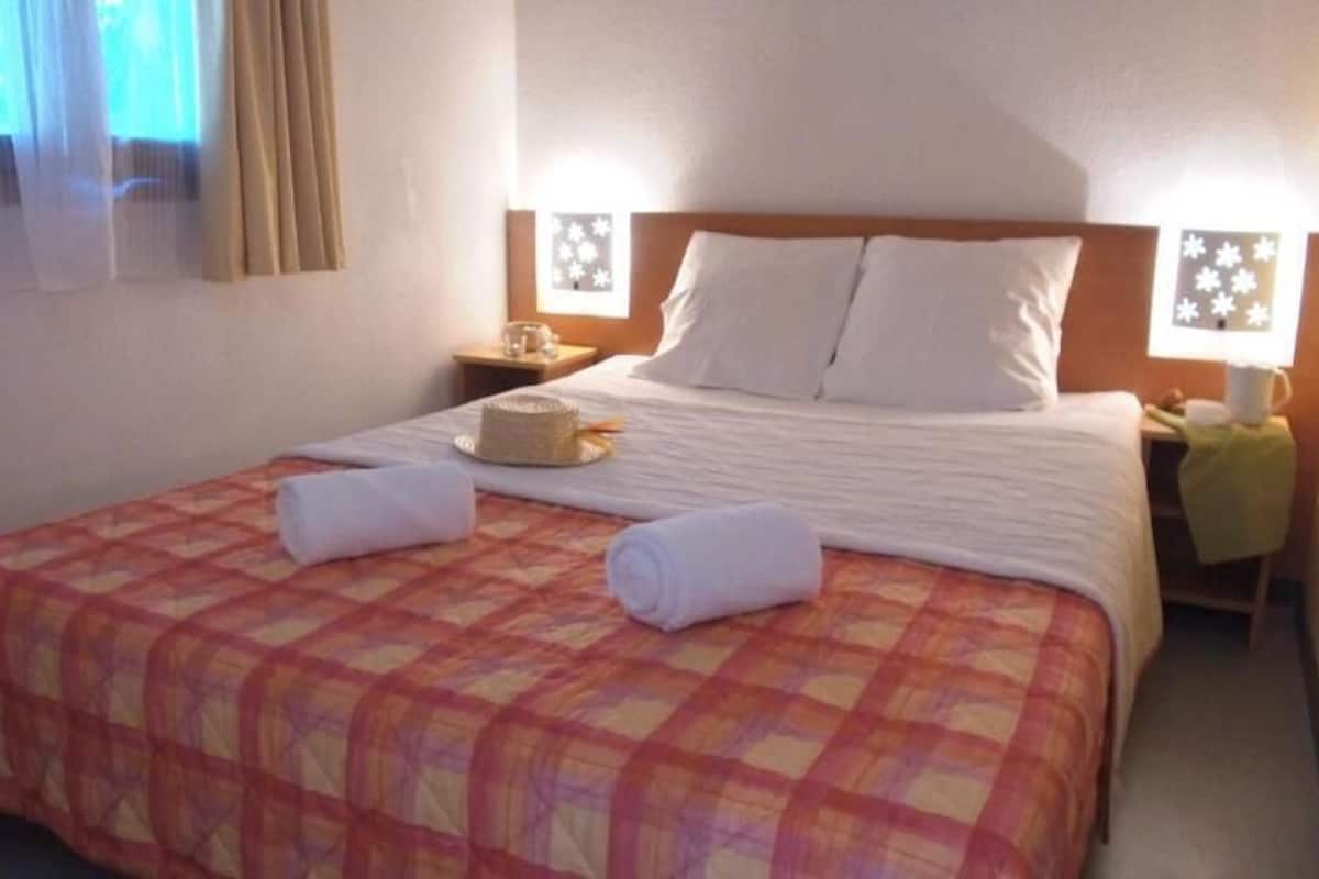 Quarto de hotel com cama, duas mesinhas de cabeceira, um criado-mudo, janela com cortinas e luminária na parede.