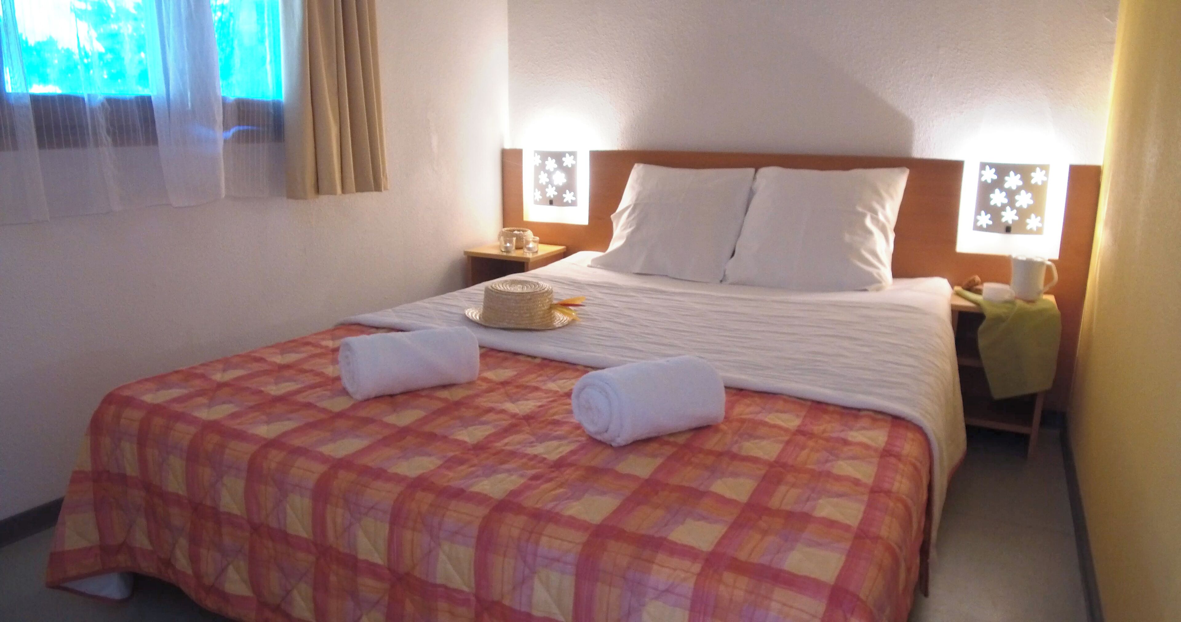 Een hotelkamer met een bed, kussens, een nachtkastje, een lamp en een raam met gordijnen.