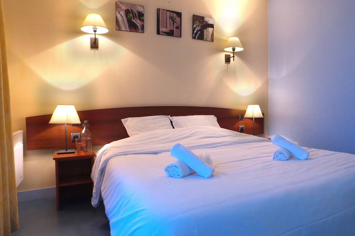 A neatly made bed with white linens and two rolled towels, two bedside lamps, and three framed pictures on the wall.