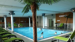 An indoor swimming pool with multiple lanes, surrounded by green lounge chairs and a palm tree.