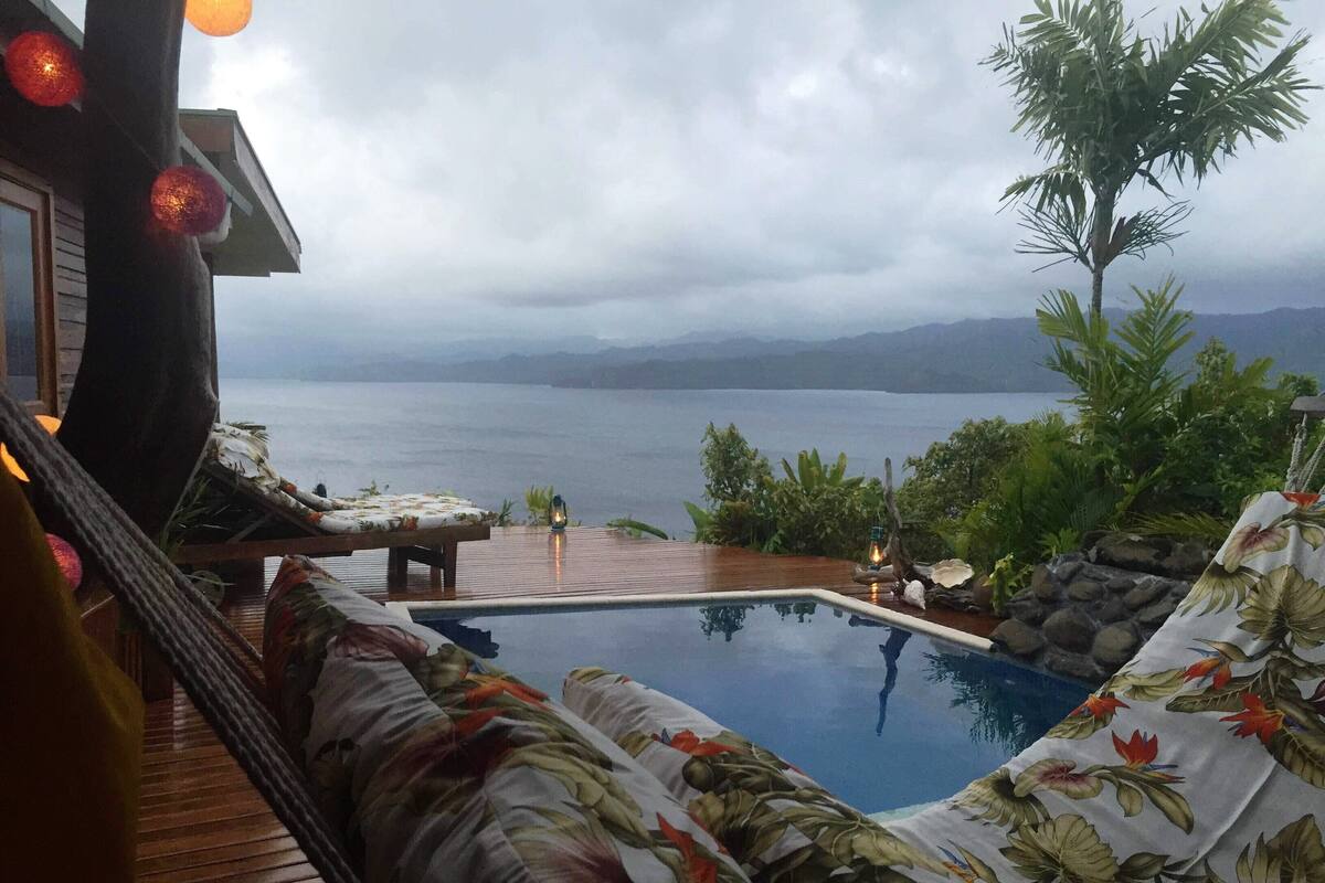 A poolside area with a wooden deck, a lounge chair, and a view of the sea and mountains.