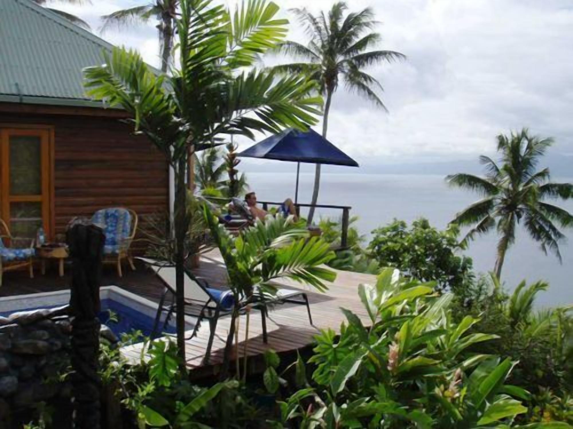 A wooden cabin with a deck overlooking a body of water, surrounded by tropical vegetation.