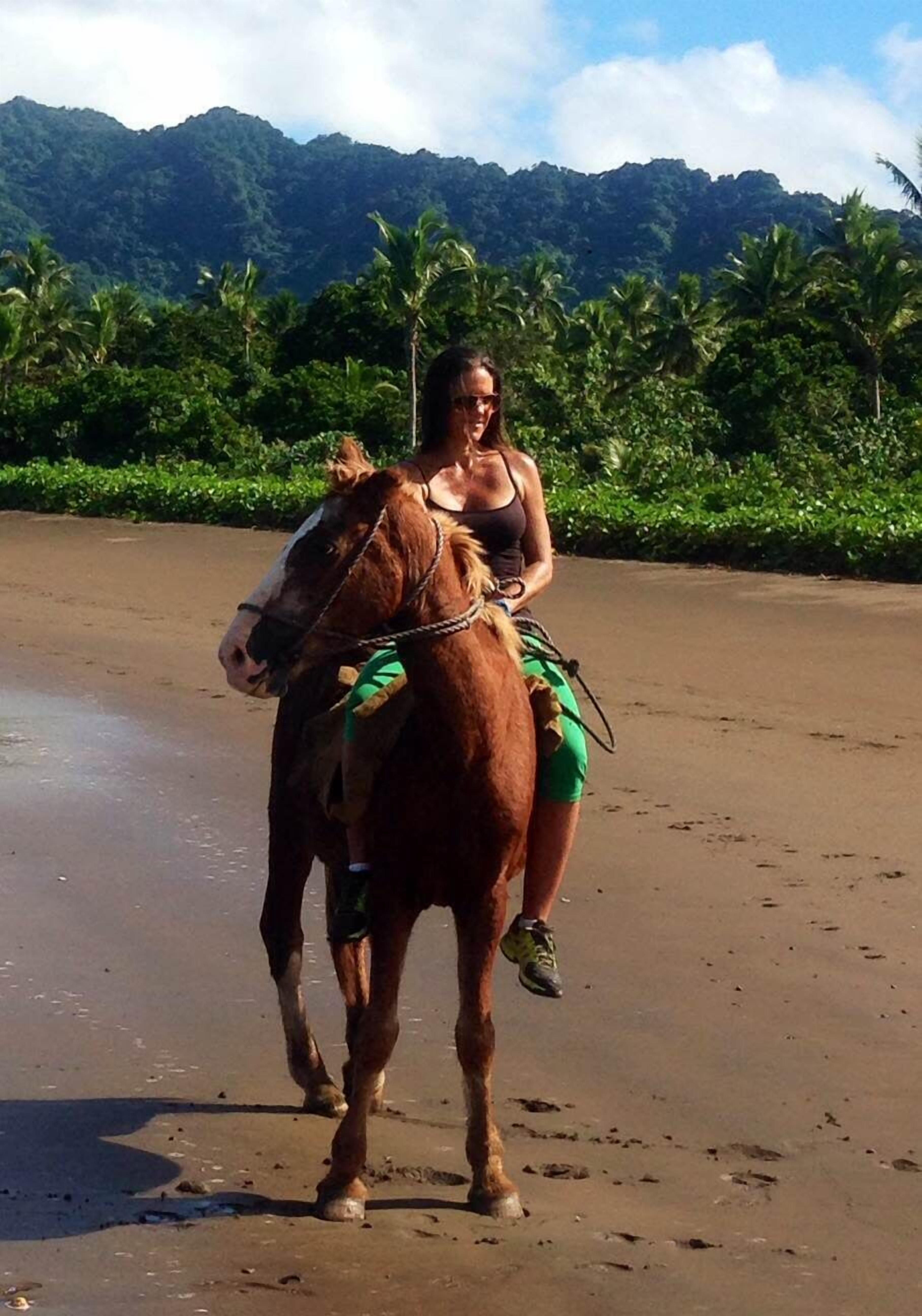 Una persona monta a caballo en una playa arenosa con abundante vegetación y montañas al fondo.
