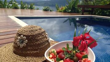 A wooden deck with a woven hat and a bowl of fruit salad by a pool with a view of mountains.