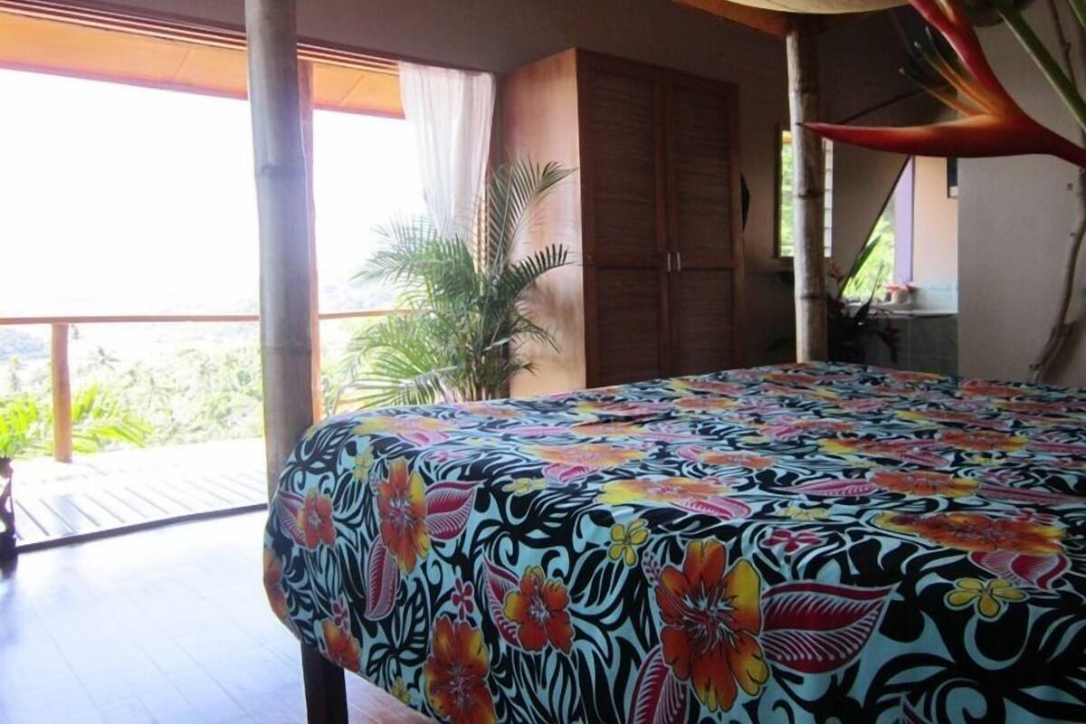 A room with a bed covered in a floral-patterned bedspread, a wooden wardrobe, and a view of greenery outside.