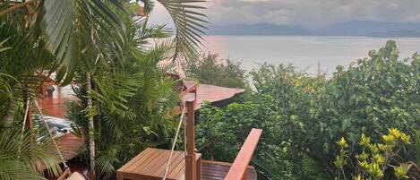 Um deck de madeira com uma cadeira de balanço e uma mesa, com vista para um corpo de água e montanhas.