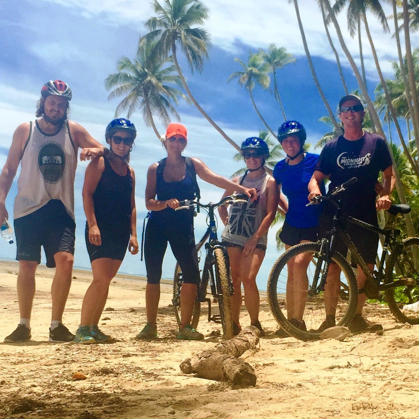 A group of people on a sandy beach with bicycles, palm trees, and a clear sky.