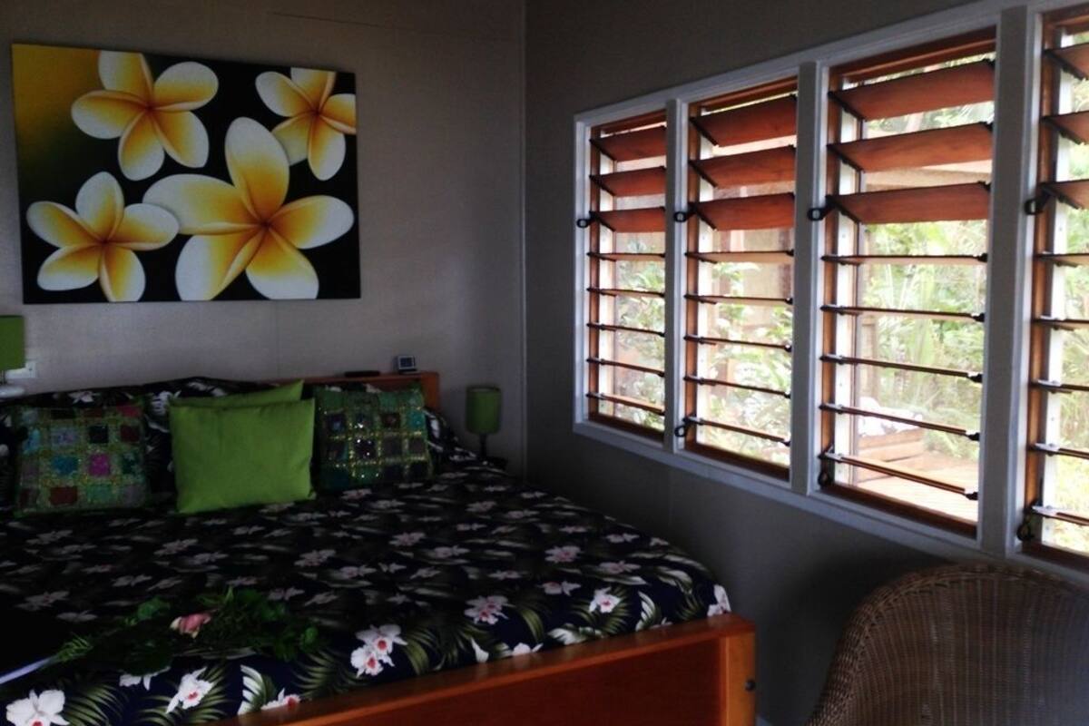 A bedroom with a bed, pillows, a green lamp, a wicker chair, and a window with wooden shutters.