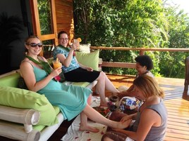 Un grupo de personas sentadas en una terraza de madera con cojines verdes, disfrutando de bebidas y aperitivos.