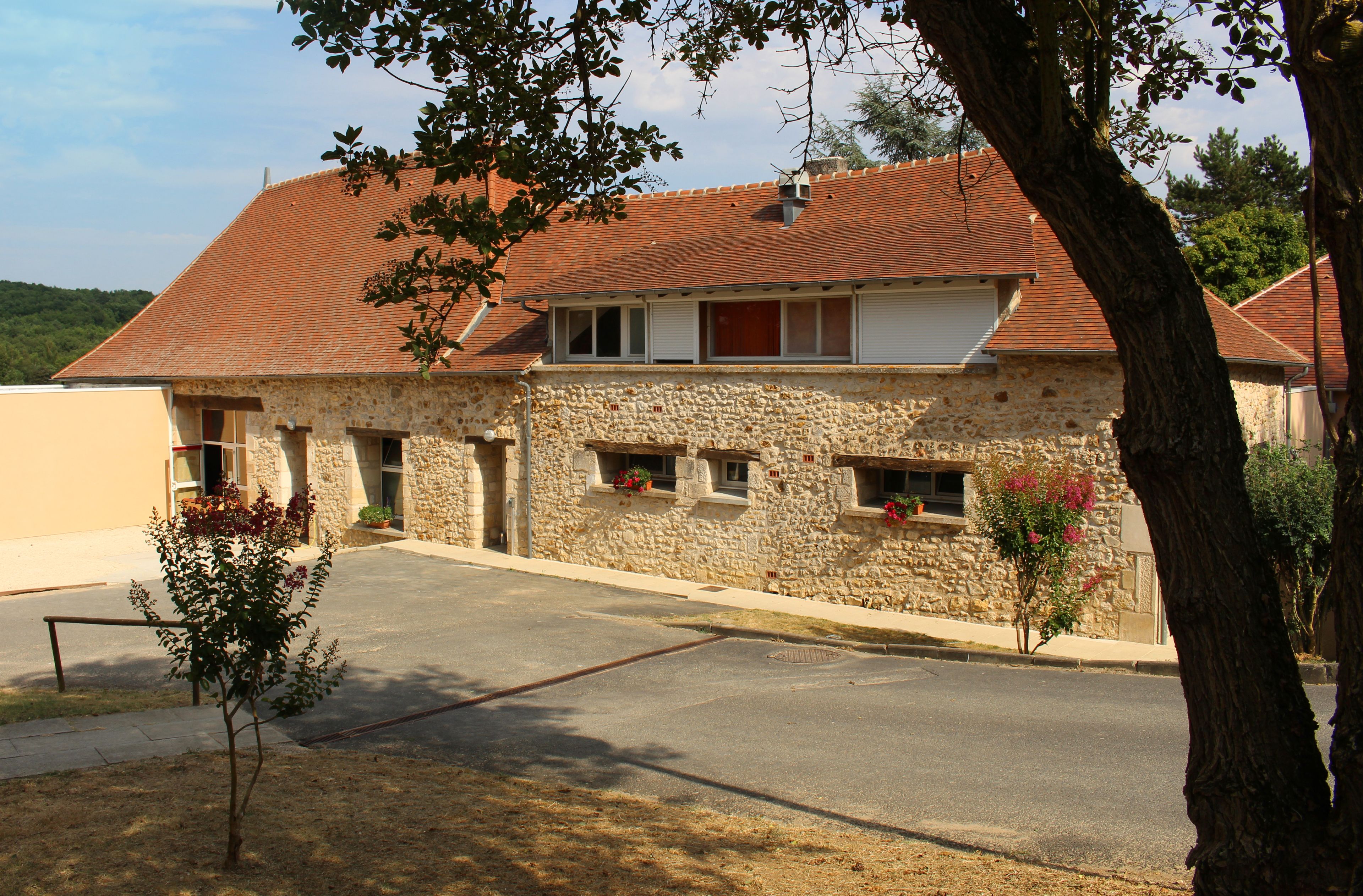 A two-story stone building with a red-tiled roof, a small front garden, and a paved driveway.
