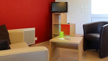Una habitación con una pared roja, un televisor sobre un mueble, una mesa con una revista, una silla y un radiador.