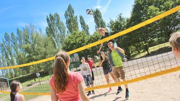 A group of people playing volleyball on a sandy court with a yellow net, surrounded by trees.