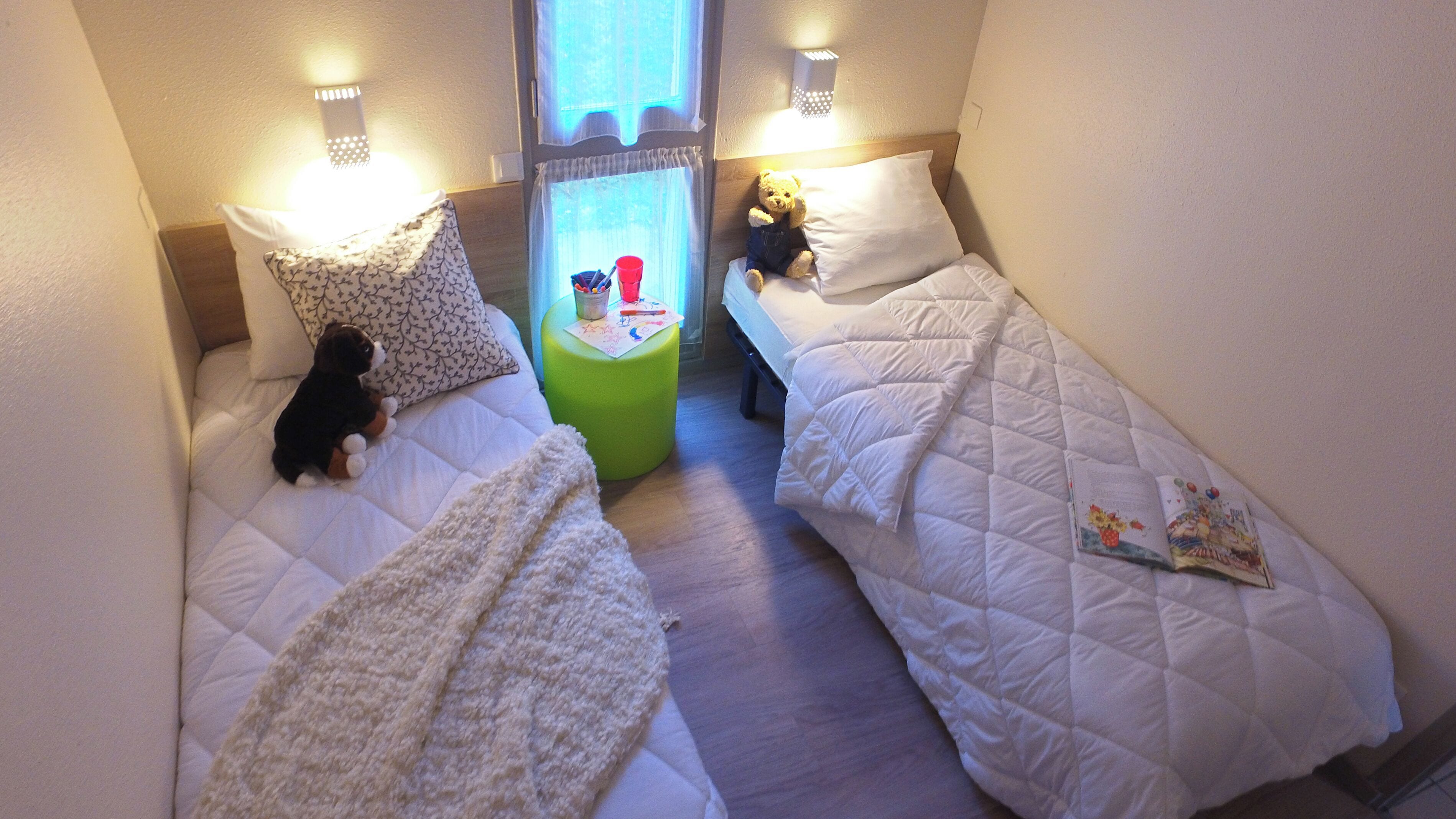 A small, cozy room with two beds, a green stool, and a teddy bear on one bed.