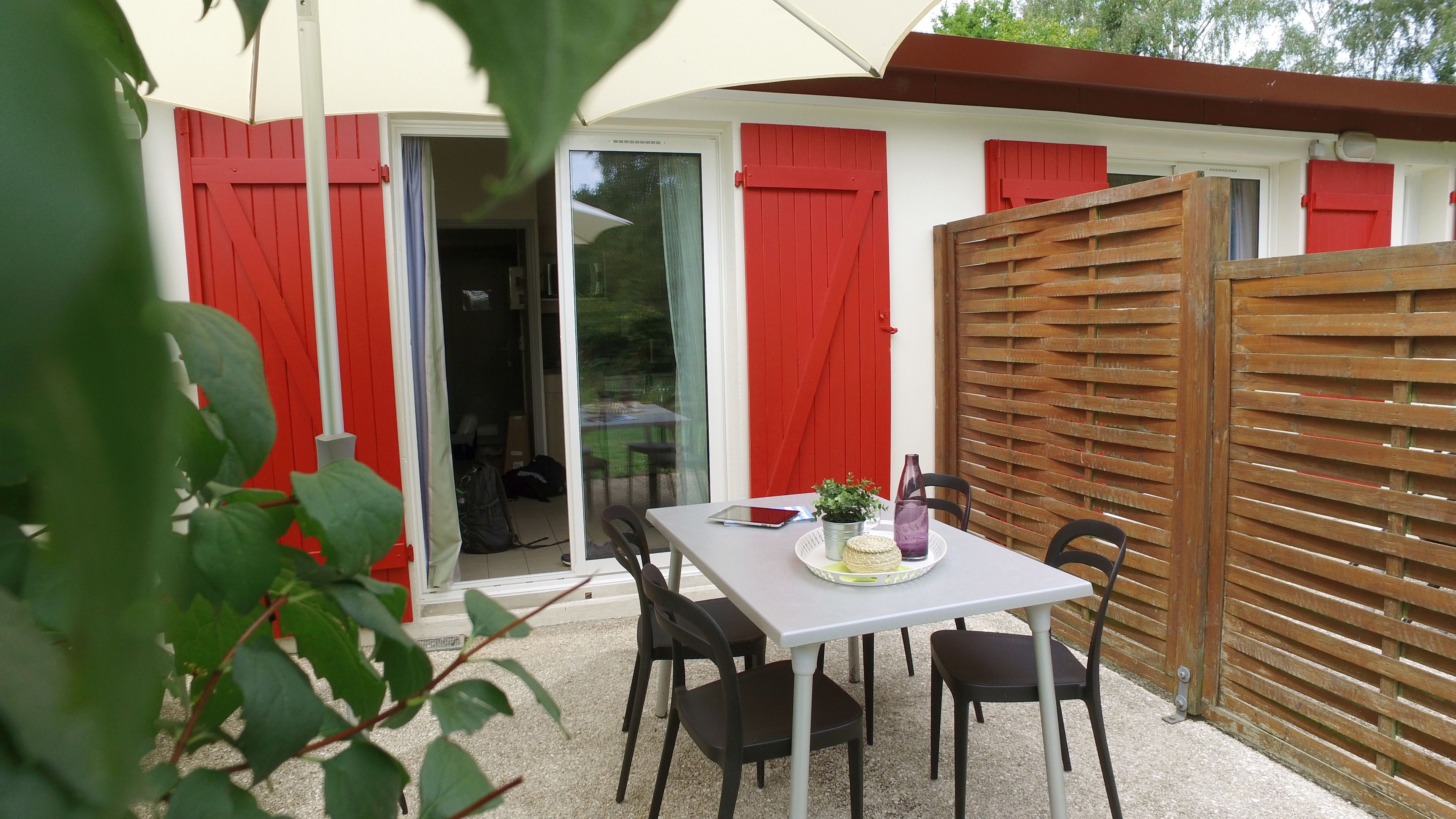 An outdoor patio with a table and chairs, red shutters, and a wooden fence.