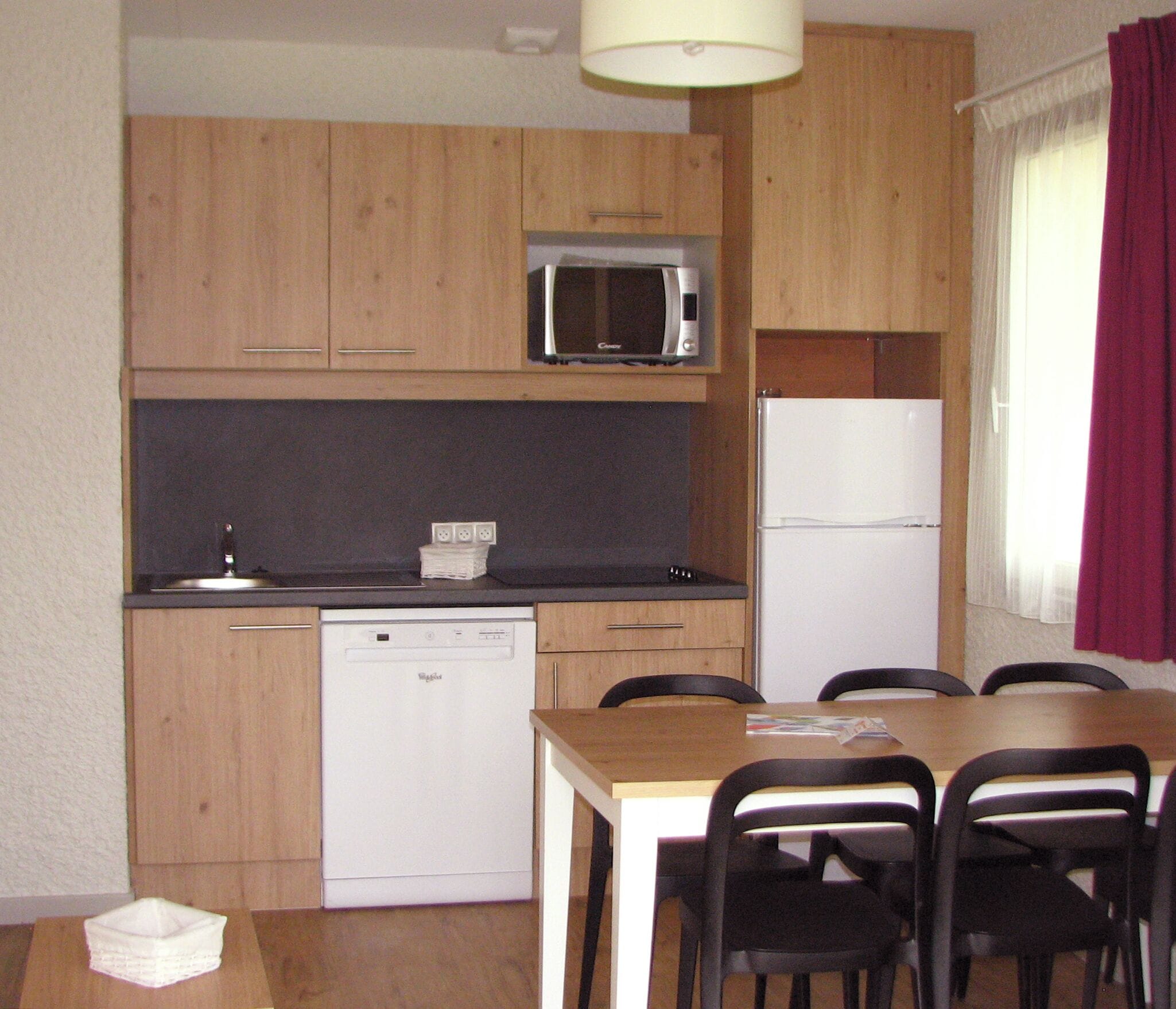 Une cuisine compacte avec des meubles en bois, un four à micro-ondes, un lave-vaisselle et un réfrigérateur. Au centre, on trouve une table à manger avec des chaises.