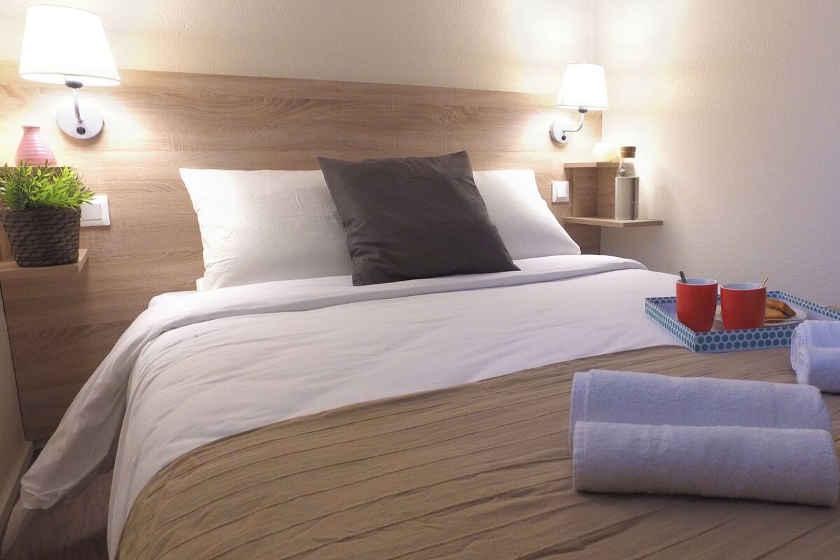 A neatly made bed with white and dark pillows, a wooden headboard, bedside lamps, a small plant, a tray with red cups and a bottle, and rolled towels on the bed.