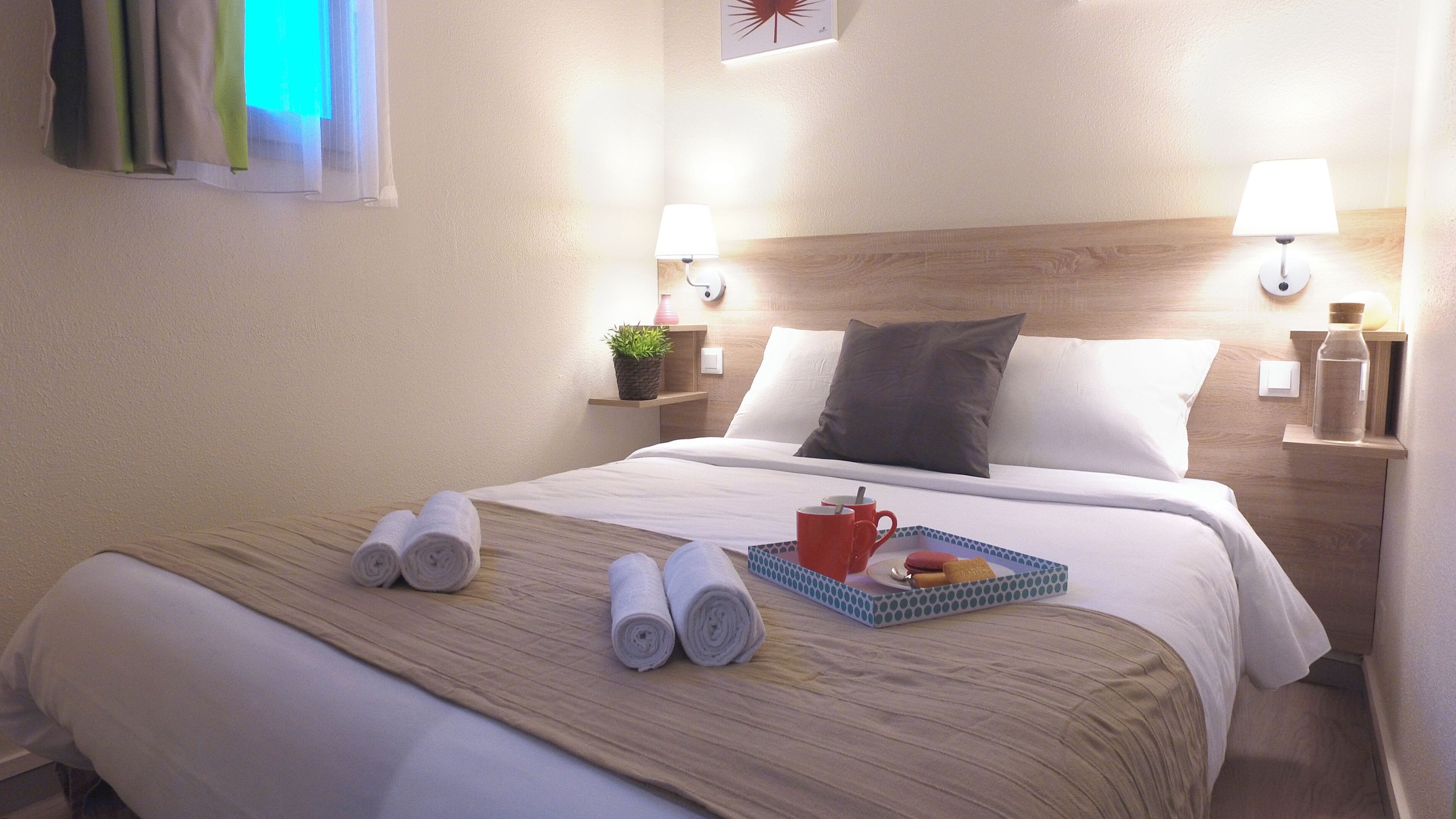 A neatly made bed with rolled towels, a tray with a mug and snacks, and two wall-mounted lamps.