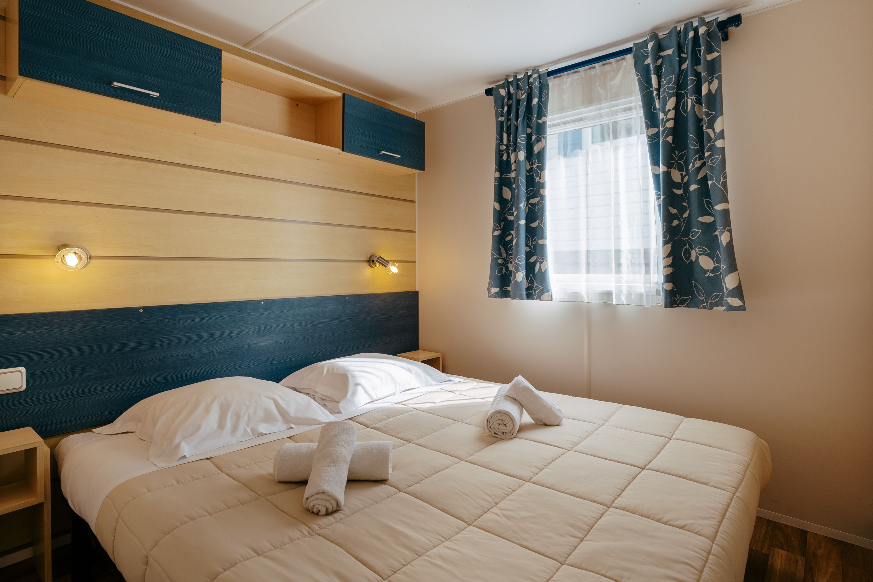 Een netjes opgemaakt bed met een gewatteerde spreideken, twee nachtkastjes en een raam met bloemen gordijnen.
