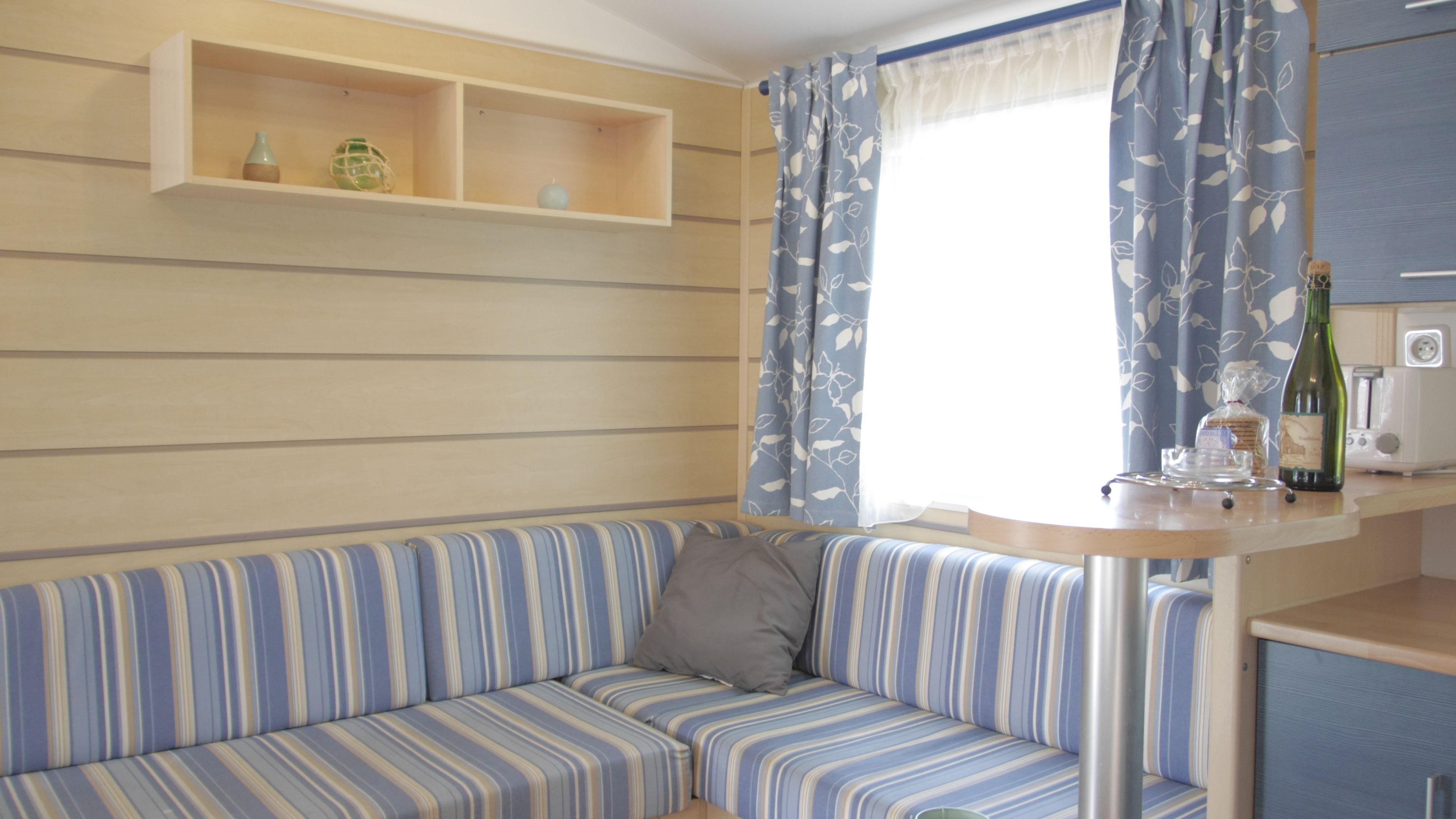 A striped sofa with a cushion, a small table with a bottle and glasses, and a window with curtains.